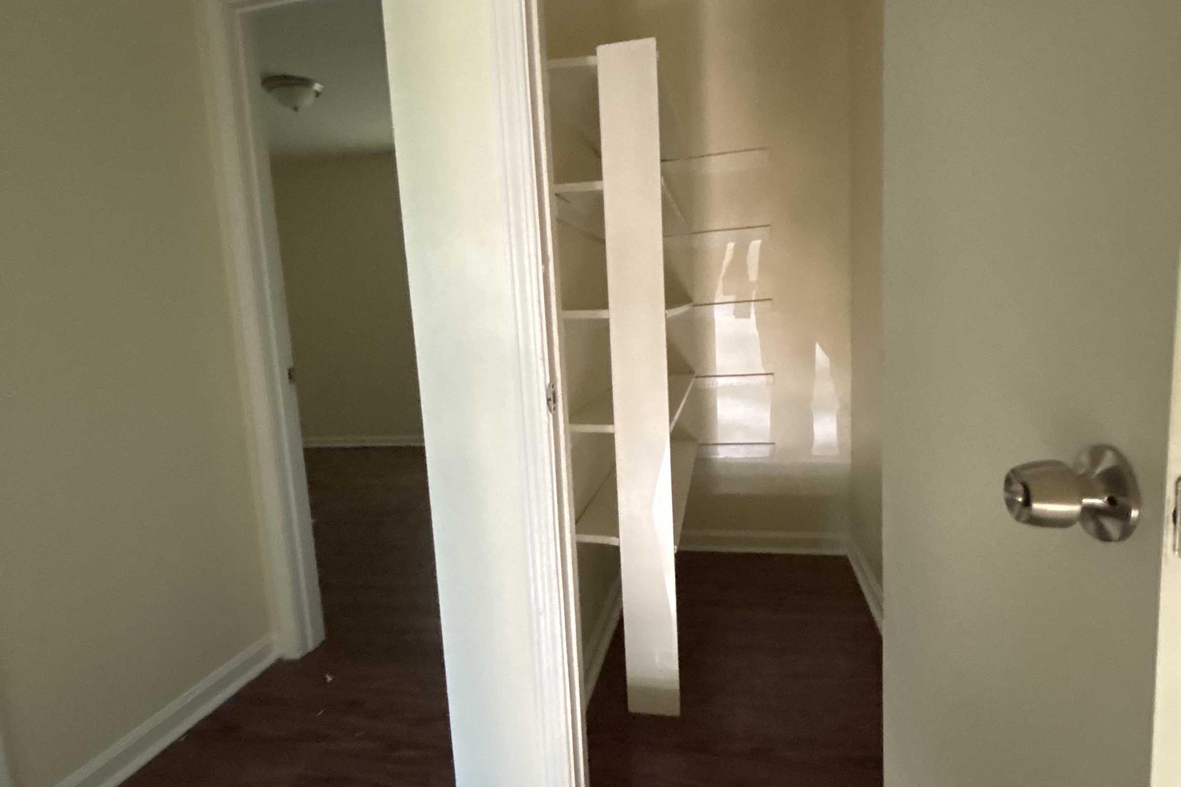 A view of a narrow room with light-colored walls. There is a wooden floor and a door slightly ajar, revealing a shelving unit against one wall. The room appears empty and well-lit, with a hint of another room visible in the background.
