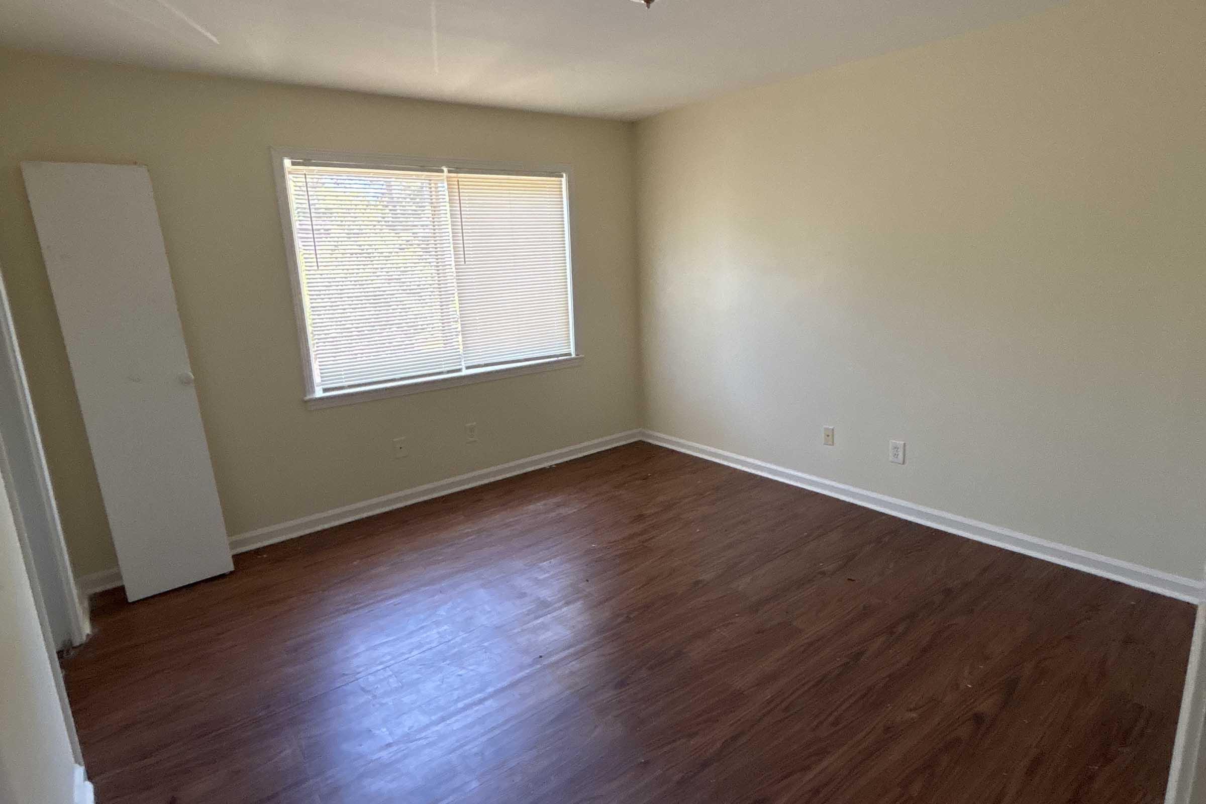 A vacant, well-lit room with light-colored walls and a hardwood floor. There's a window with blinds providing natural light, and a white wardrobe or cabinet against one wall. The overall space appears clean and empty, making it suitable for various uses.