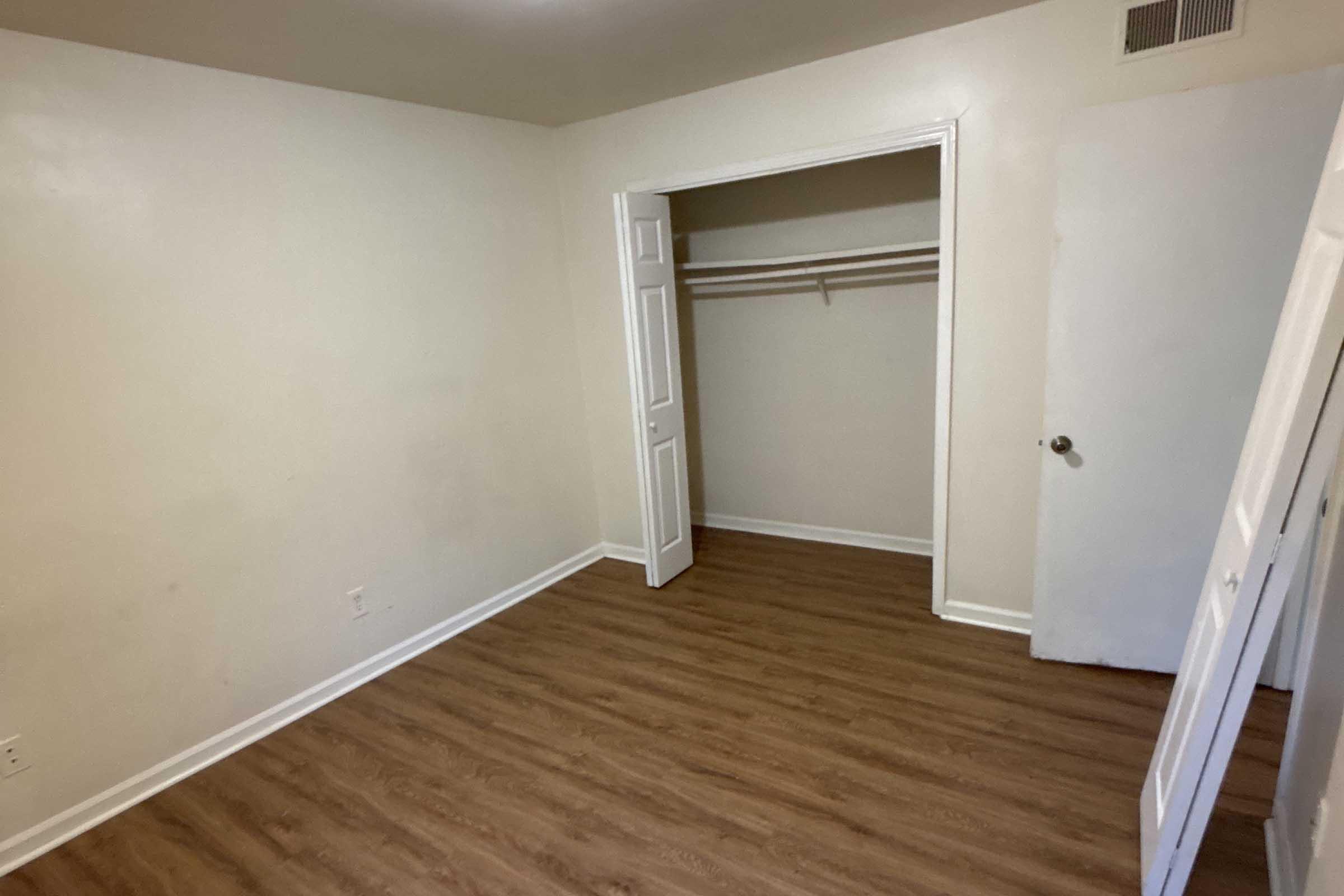 A small, empty room with light-colored walls and wooden laminate flooring. There is a closet with a hanging rod on one side and a door leading to another area. The room features a simple, minimalistic design without furniture or decorations.