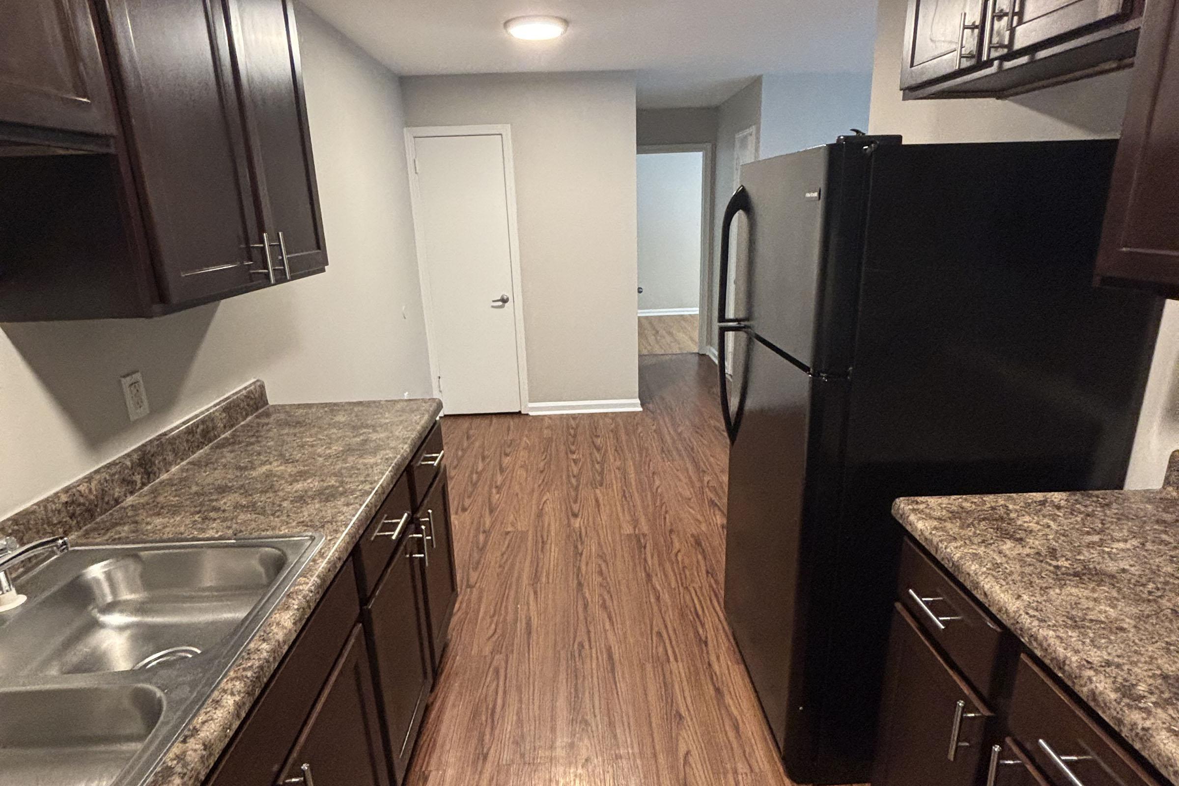 View of a modern kitchen featuring dark wood cabinets, a large black refrigerator, and a countertop with a sink. The flooring is a rich brown wood-like laminate. A hallway leads to another room in the background, with a plain wall and door visible. The space is well-lit and tidy.