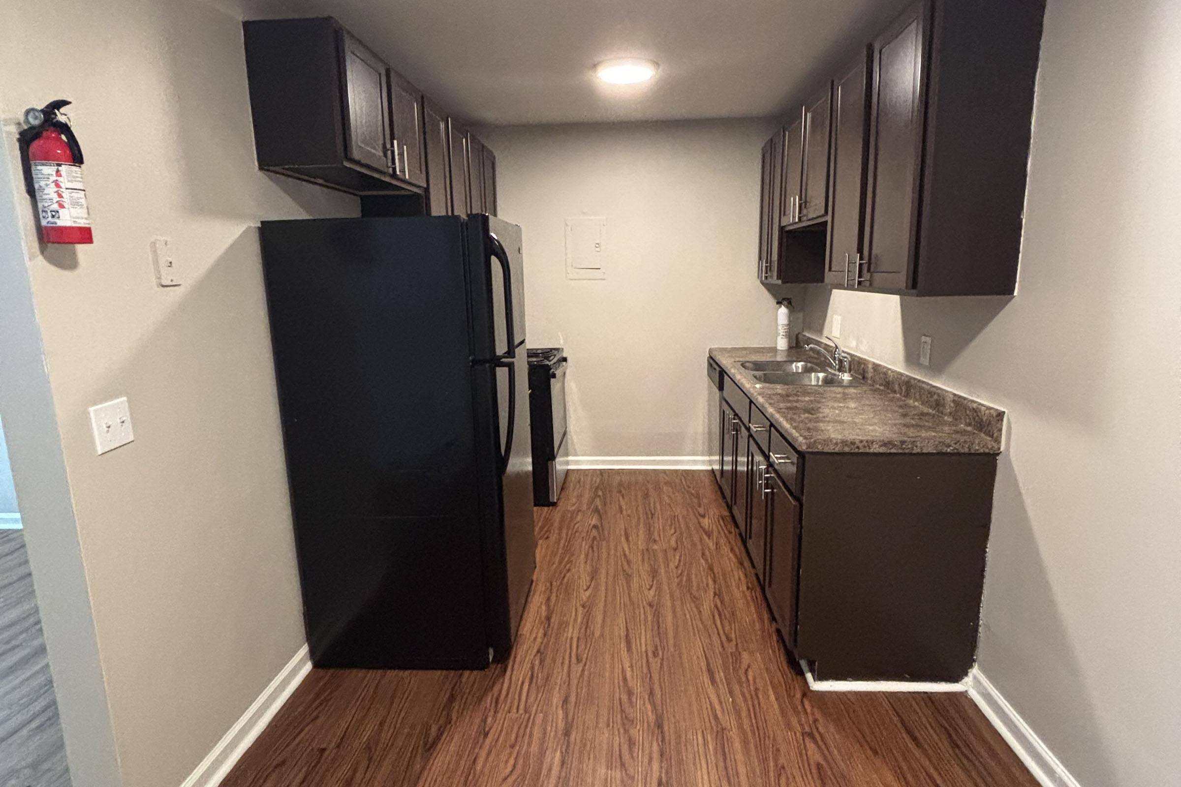 A small kitchen featuring dark wood cabinets, a black refrigerator, and a black stove. The countertop is made of granite, and the flooring is laminate wood. A fire extinguisher is mounted on the wall near the entrance. The kitchen is well-lit with overhead lighting.