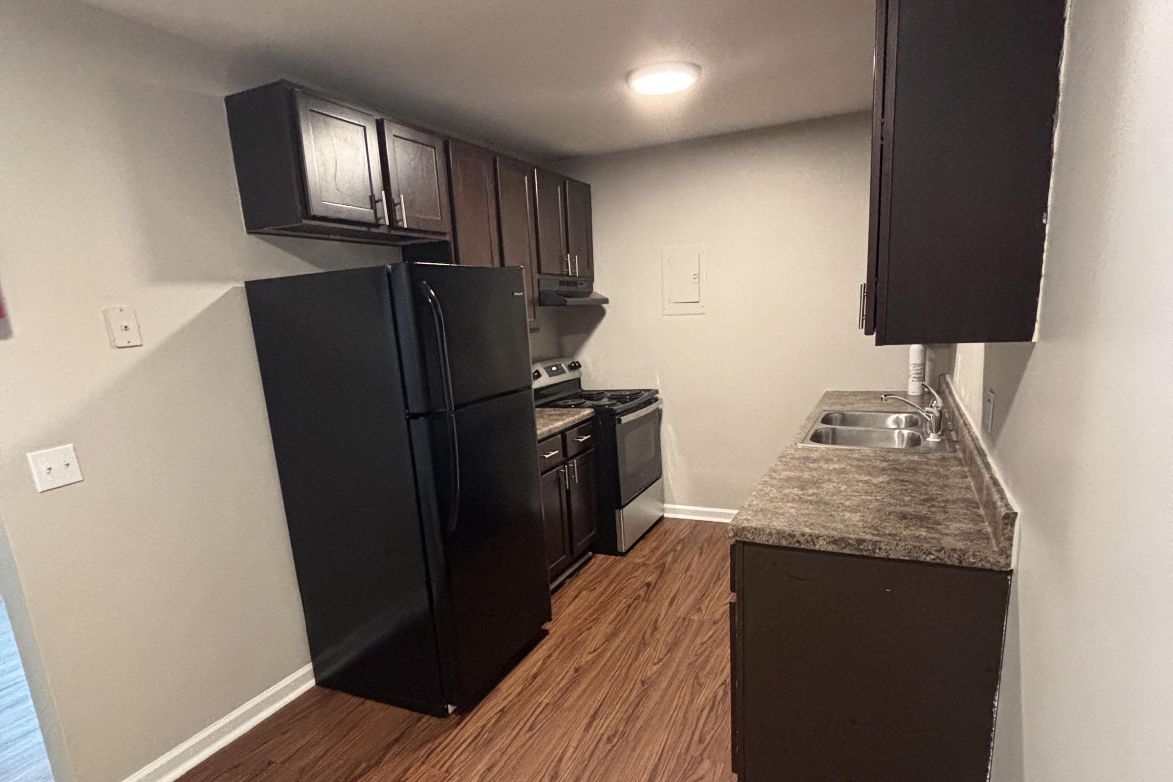 A modern kitchen featuring dark wood cabinets, a black refrigerator, a silver stove, and a countertop with a sink. The floor is made of dark laminate wood. The walls are painted light gray, and the kitchen is well-lit with overhead lighting.