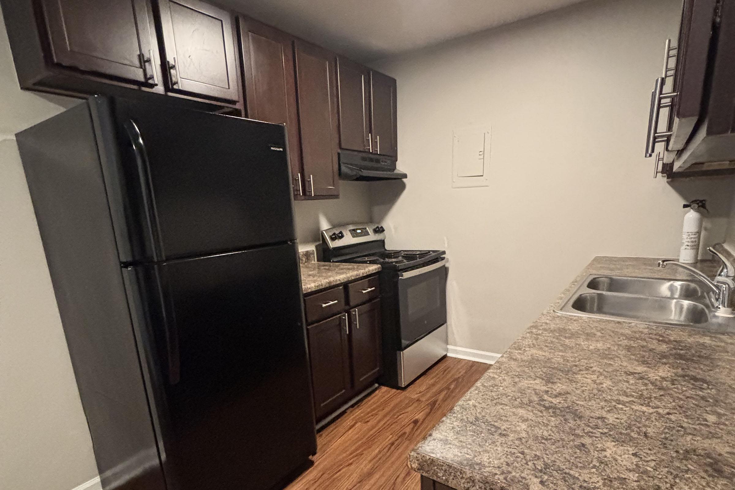 A modern kitchen featuring dark wooden cabinets, a black refrigerator, a stainless steel stove, and an overhead microwave. The counter is made of a granite-like material, and there are two stainless steel sinks. The walls are light-colored, and the floor has a wooden finish.