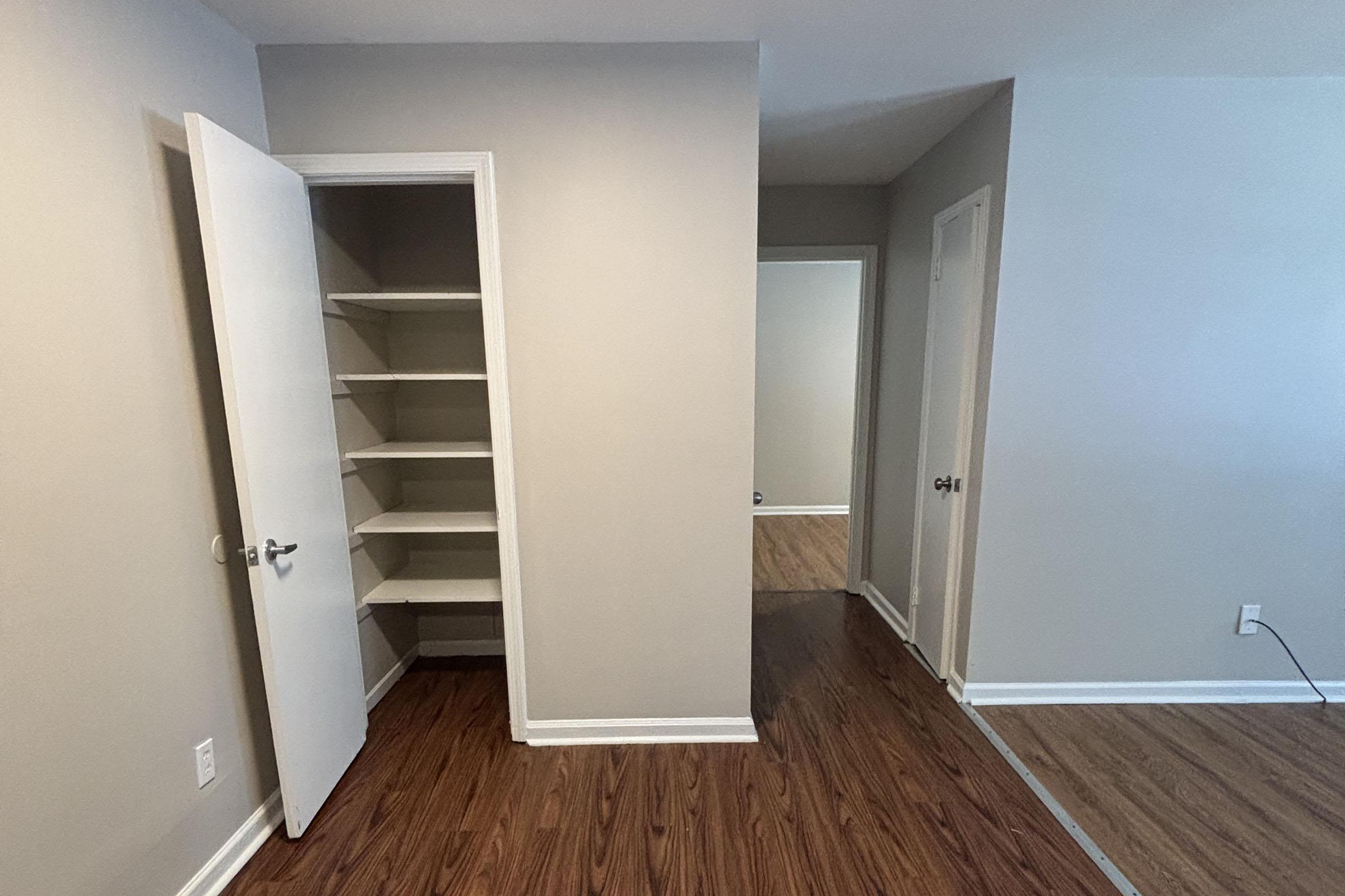 A view of a room with light-colored walls and wooden laminate flooring. There is an open closet with shelves on the left side, and a doorway leading to another room visible in the background on the right. The space appears clean and well-lit.