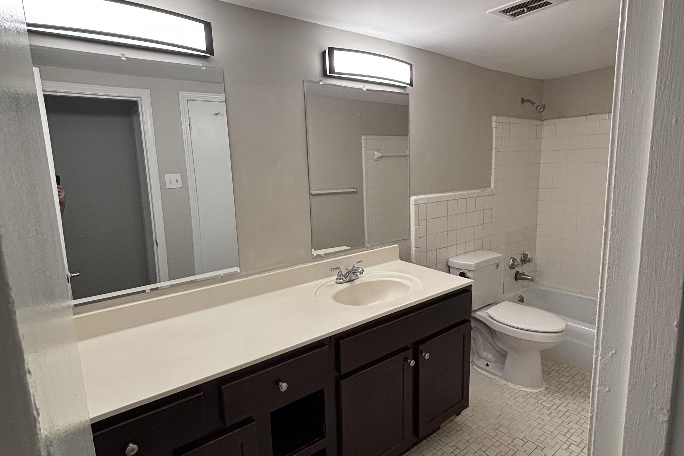 A clean, neutral-toned bathroom featuring a double sink vanity with dark cabinetry, two mirrors above the sinks, a toilet, and a bathtub with a shower. The walls are light gray, and the floor has small white tiles. There is ample natural light from an overhead fixture.