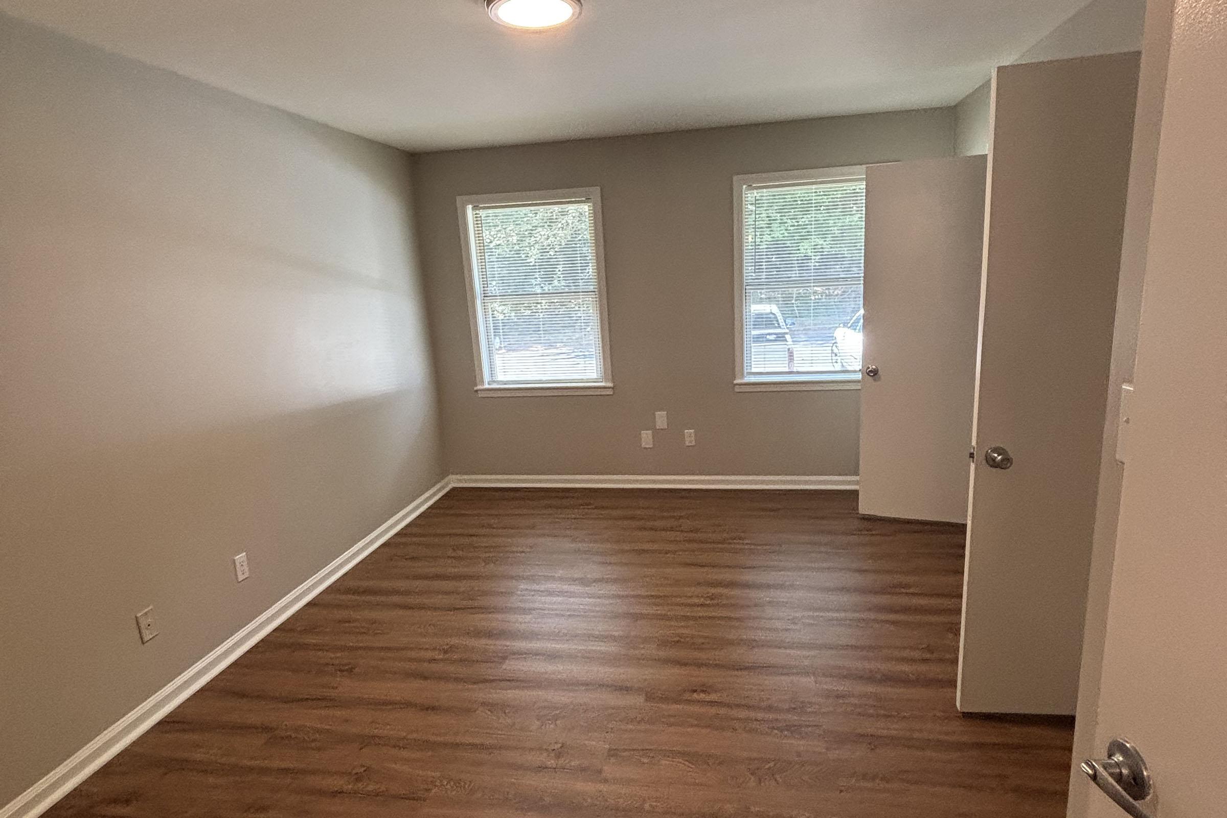Empty room with light gray walls, two windows providing natural light, and a door on the right. The flooring is made of wood-like laminate. The room has no furniture, giving it a spacious and clean appearance.