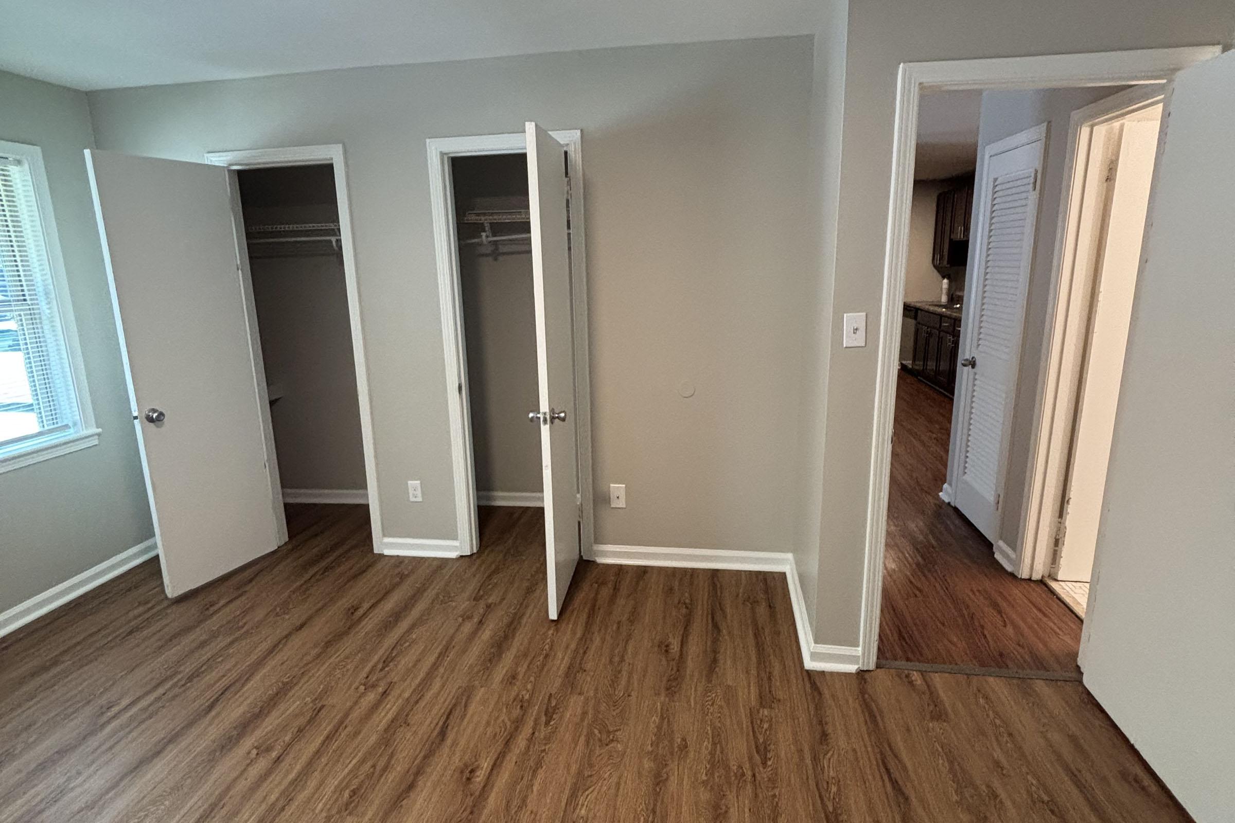 A spacious, light-colored room featuring two open closet doors on the left and a door leading to another area on the right. The floor is wooden, and there is a window on the far wall allowing natural light to enter, creating a bright and airy atmosphere.