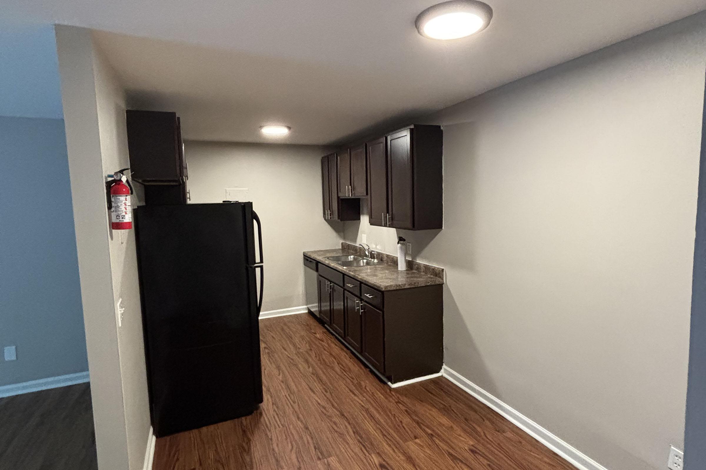 A modern kitchenette with dark wood cabinetry, a stainless steel sink, and a black refrigerator. The countertops are dark, and the space features recessed lighting. The walls are painted a light gray, and the floor has dark wood laminate. A fire extinguisher is mounted on the left wall.