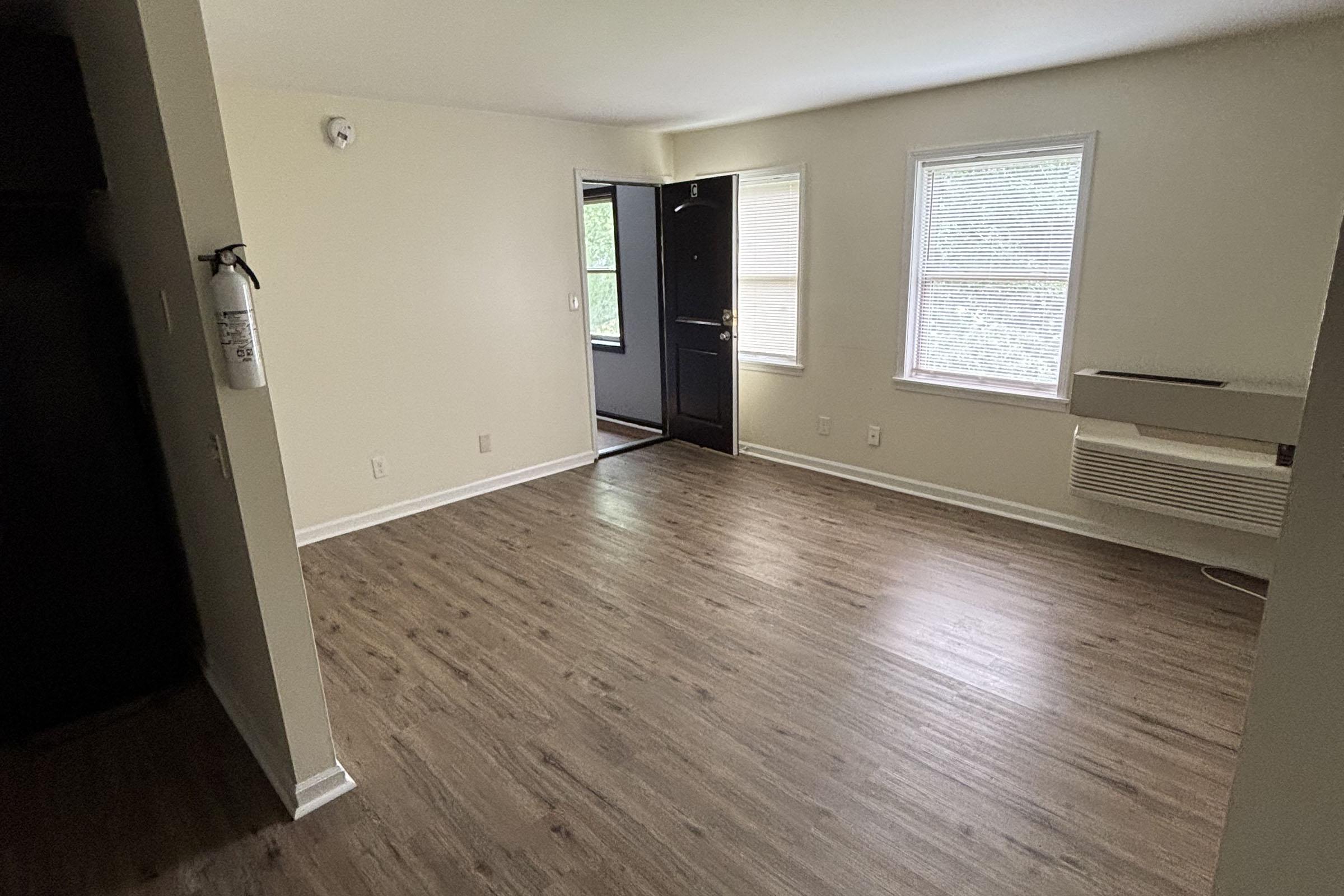 Empty living room with light-colored walls and wooden flooring. A black front door is visible in the background, leading to another room with a window. An air conditioning unit is mounted under the window. The space is well-lit and uncluttered, creating a clean and inviting atmosphere.