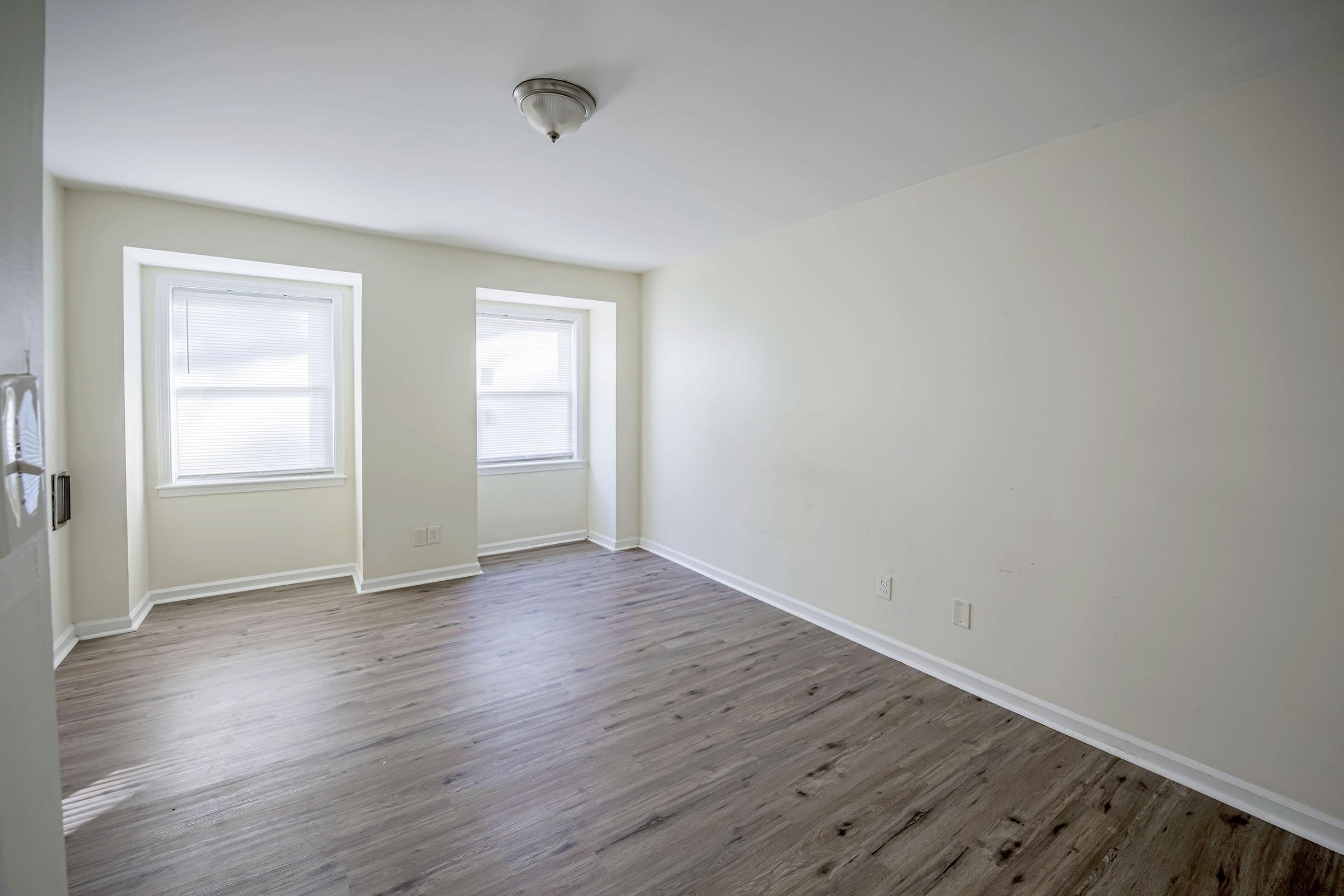 Empty room with light-colored walls and two windows letting in natural light. The floor is wooden and the ceiling has a simple light fixture. No furniture or decorations are present, creating a clean and minimalist appearance.