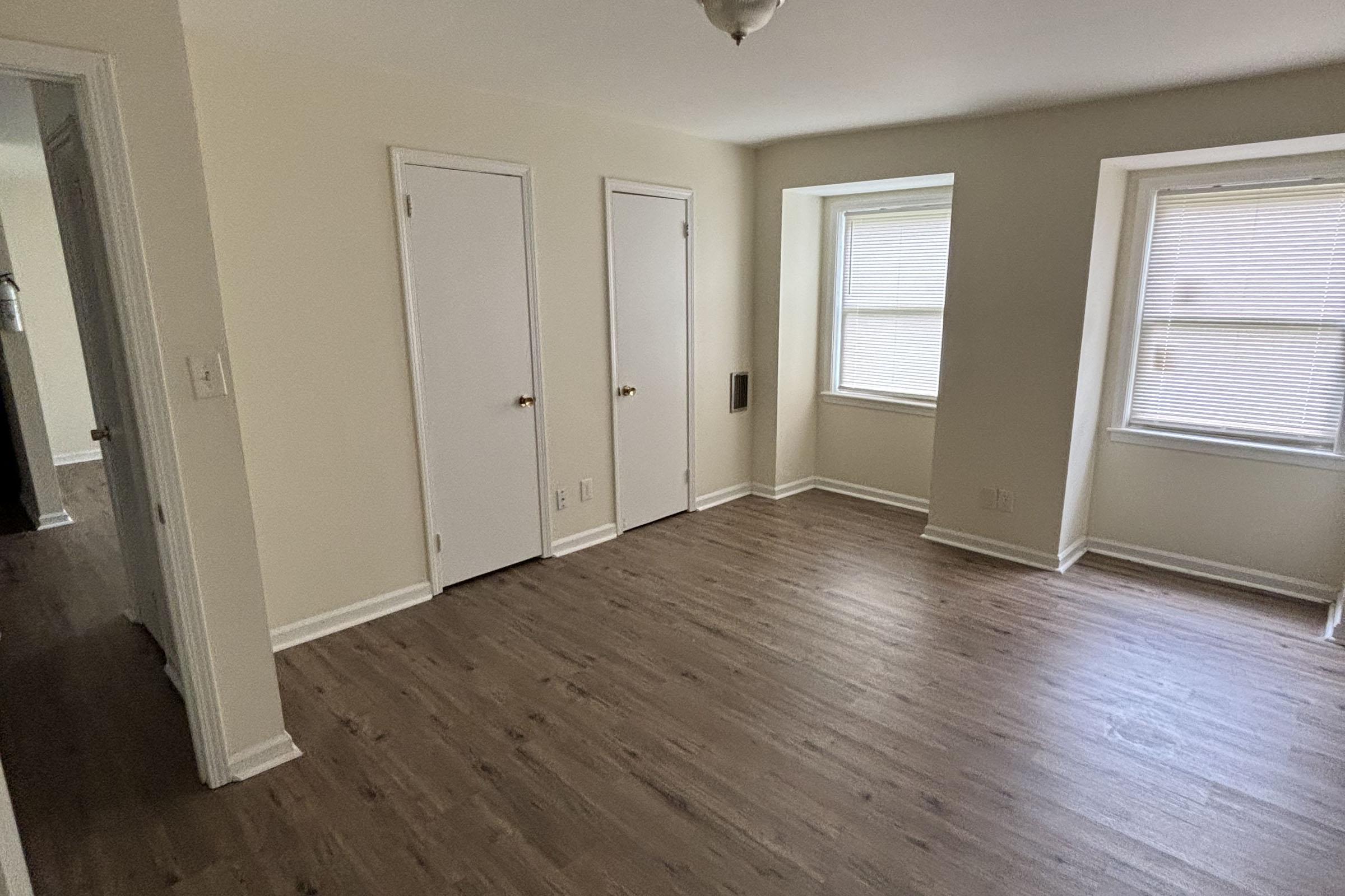 A well-lit, empty room featuring light-colored walls and laminate flooring. It has two windows with blinds, two closed white closet doors, and a ceiling light fixture. The room is clean and spacious, creating an inviting atmosphere.