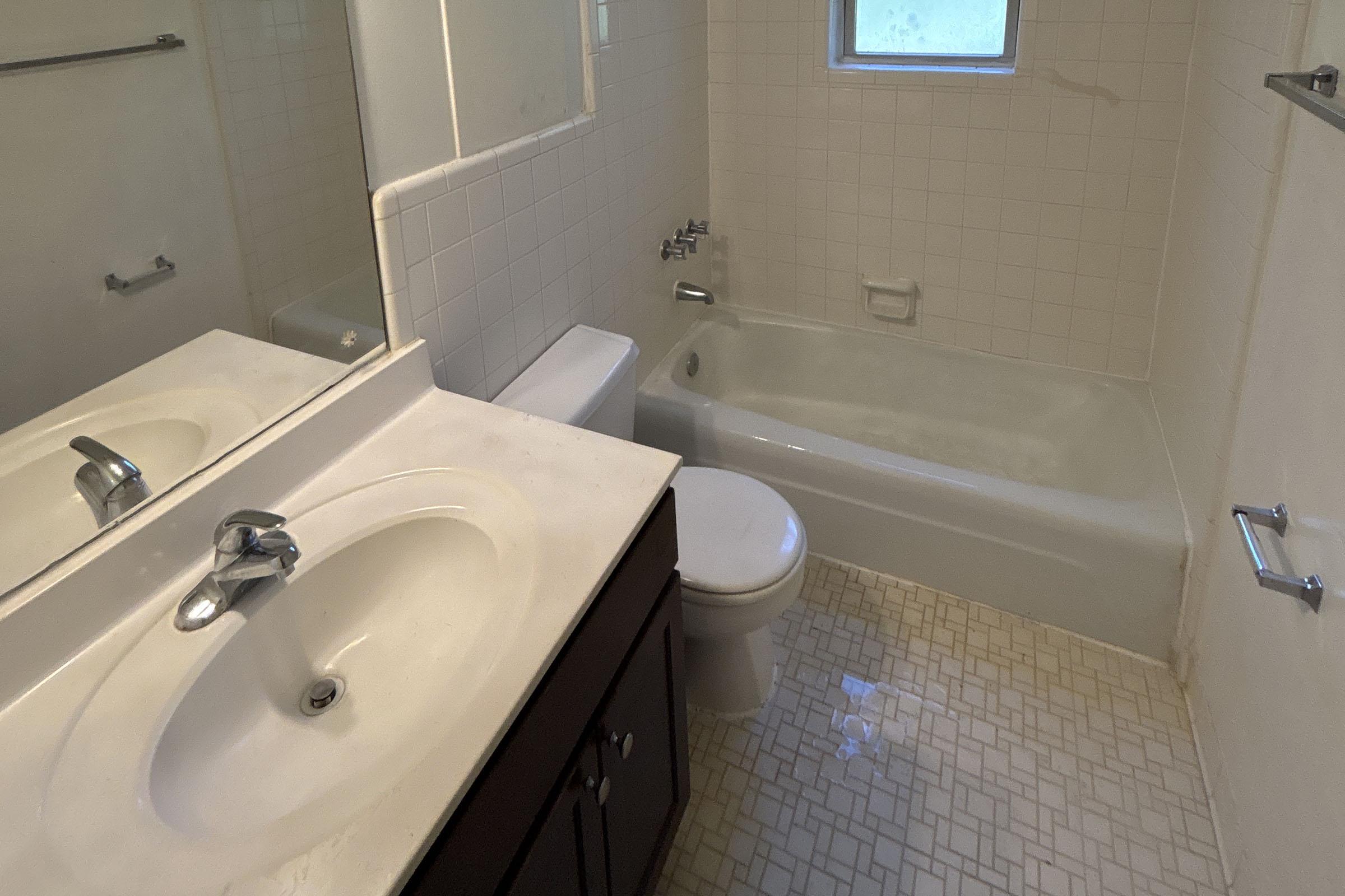 A clean, small bathroom featuring a white bathtub, toilet, and sink with a vanity. The walls are tiled, and there is a window that lets in natural light. The floor has a patterned design, and the overall decor is simple and functional.