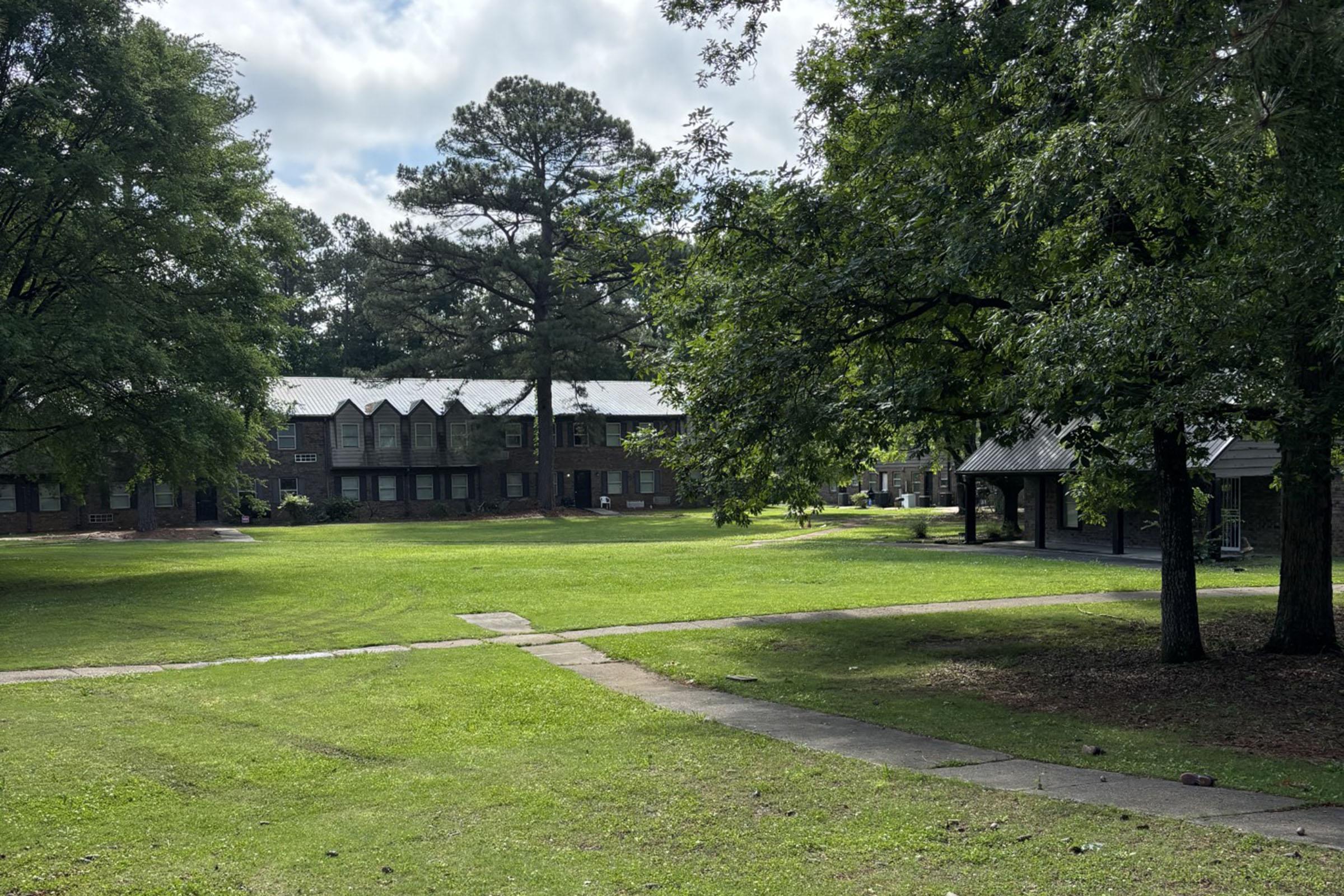 A grassy area with several large trees and two multi-story brick buildings in the background. A path leads through the grass, and there is a small pavilion nearby. The sky is partly cloudy, creating a serene outdoor environment.