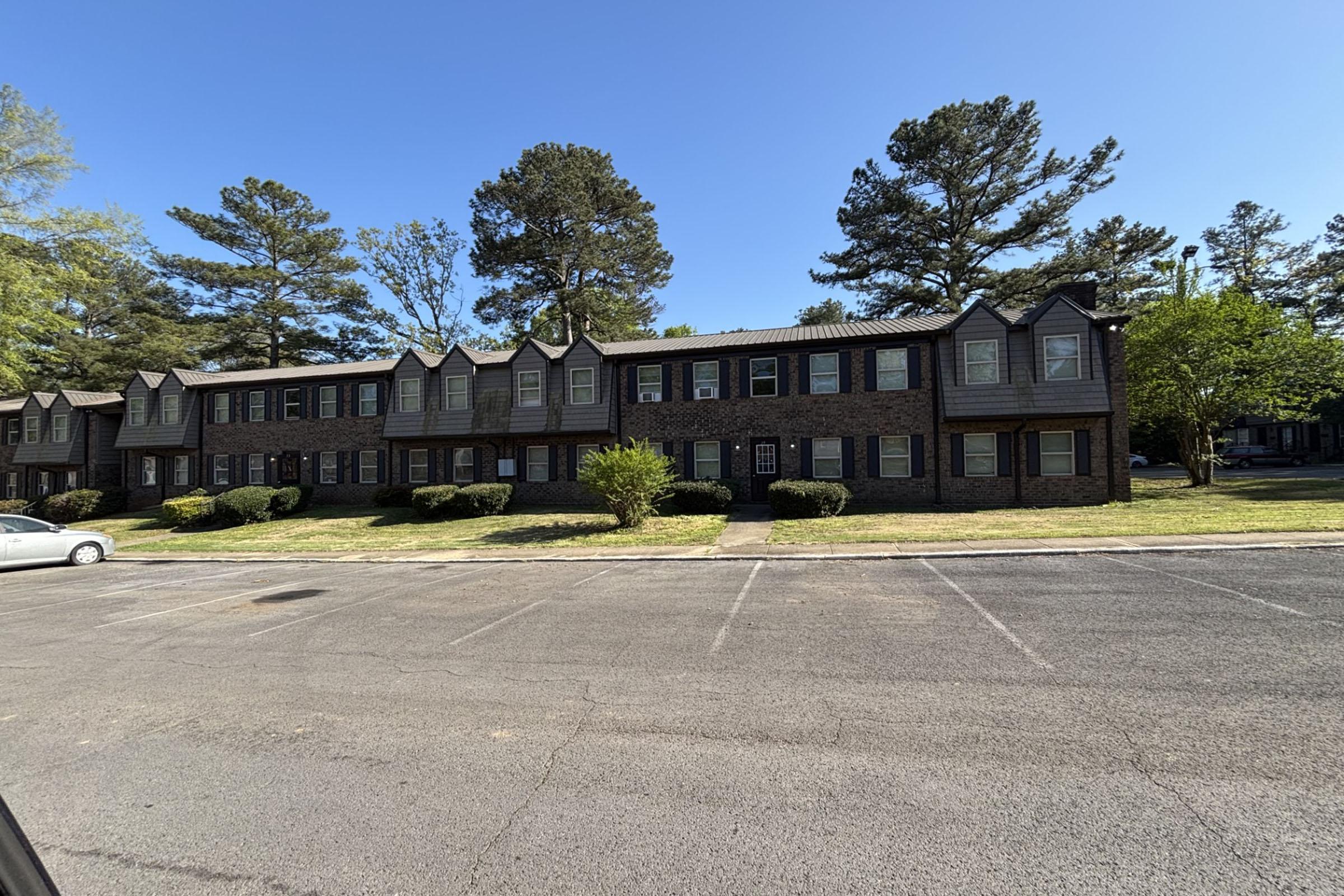 A two-story brick apartment building with multiple units, surrounded by grass and shrubs. Tall trees line the background, under a clear blue sky. A parking area is visible in front, showing several empty spaces.