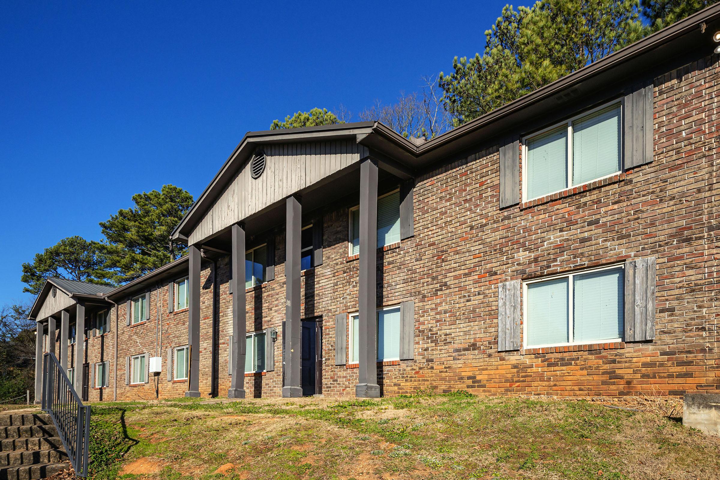 Exterior view of a two-story brick apartment building with large windows and a simple, modern entrance. The building features a classic architectural style with columns at the front, surrounded by a grassy area and tall trees in the background under a clear blue sky.