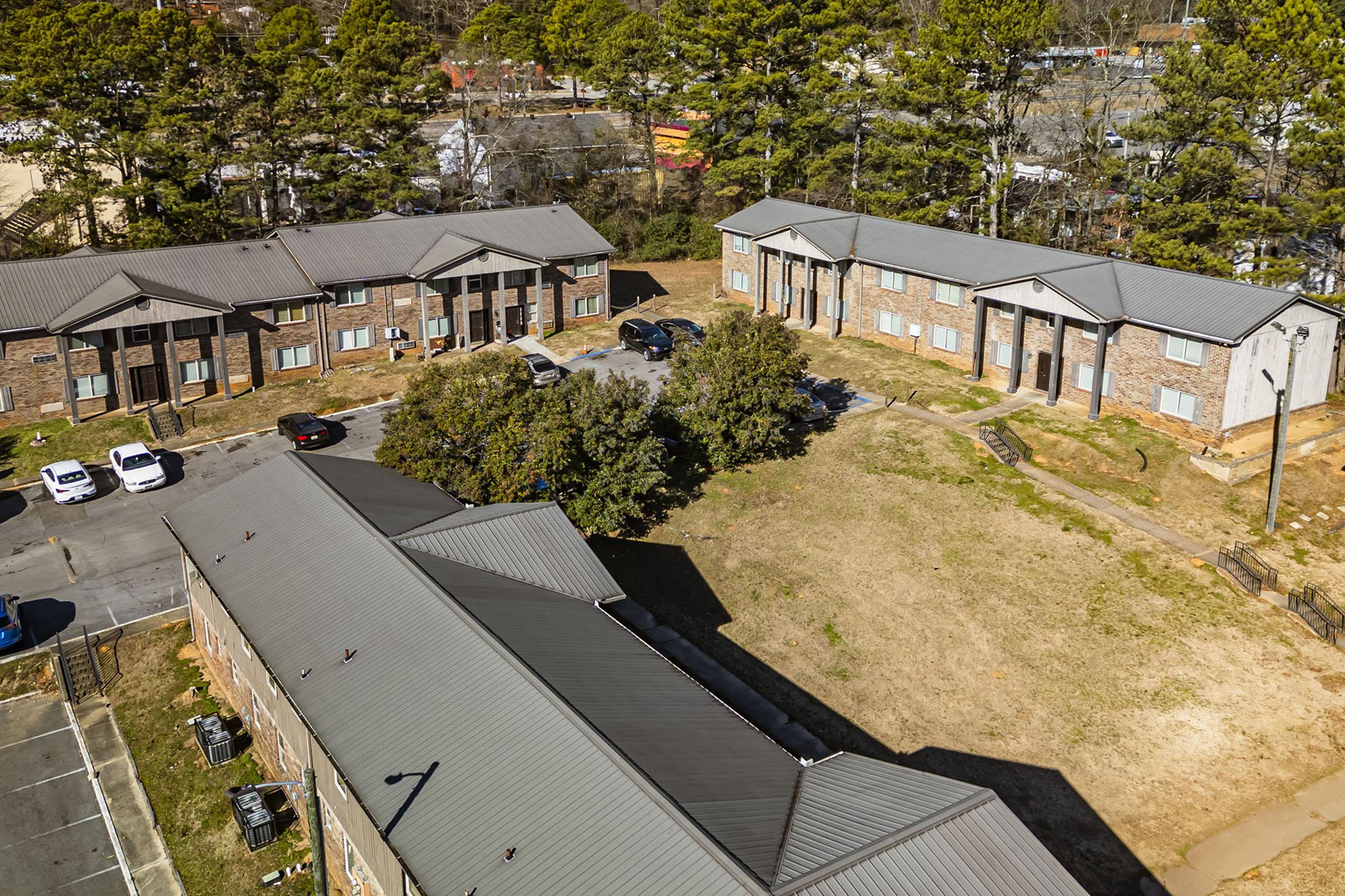 Aerial view of a residential complex featuring several buildings with brick exteriors and gray roofs. The scene shows a landscaped area with bare ground, a few parked cars, and surrounding trees. In the background, additional buildings and roadways are visible.