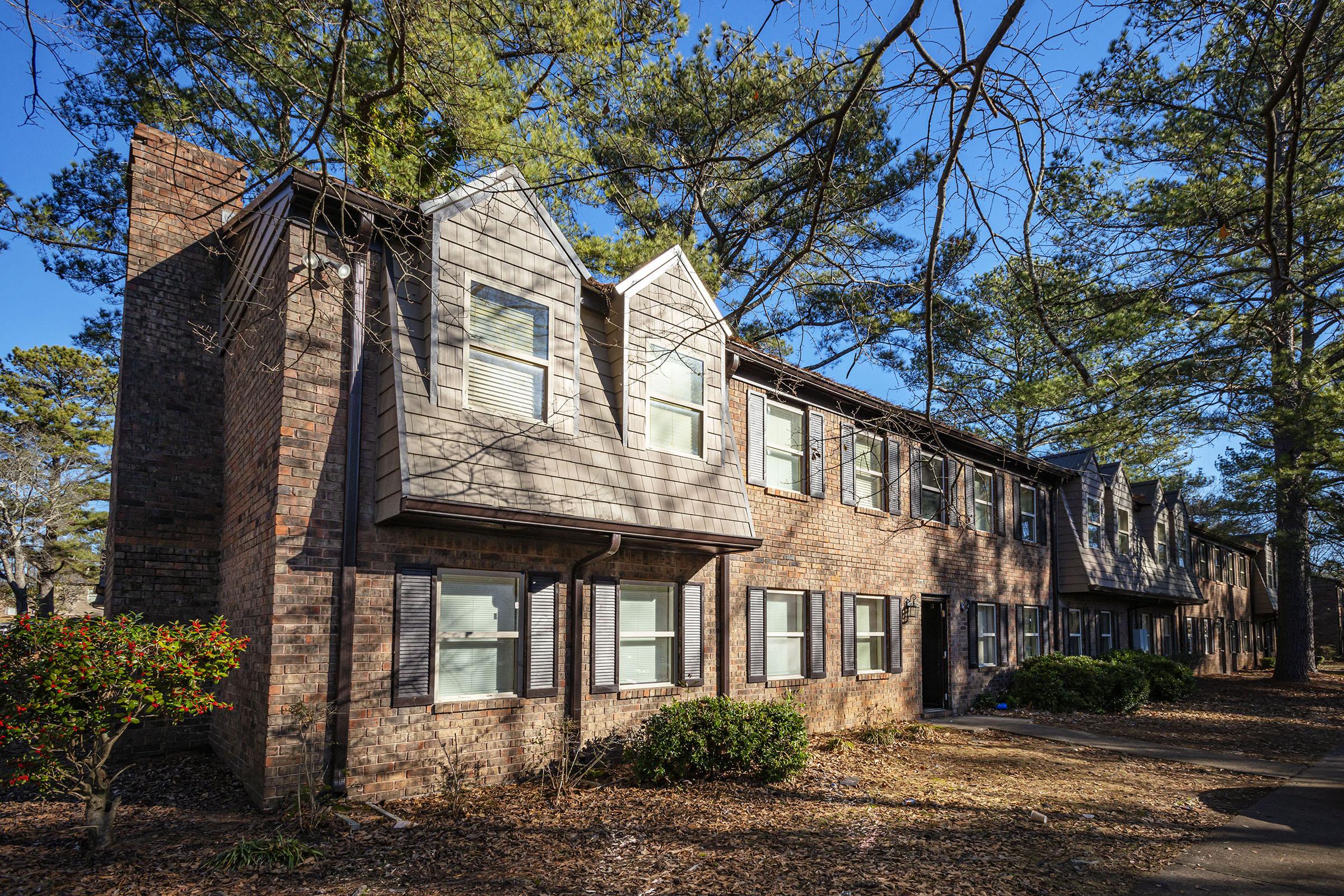 A row of brick townhouse-style buildings surrounded by trees. The houses feature peaked roofs and multiple windows, with some greenery and fallen leaves in the foreground. The scene is bathed in natural light, indicating a clear day.