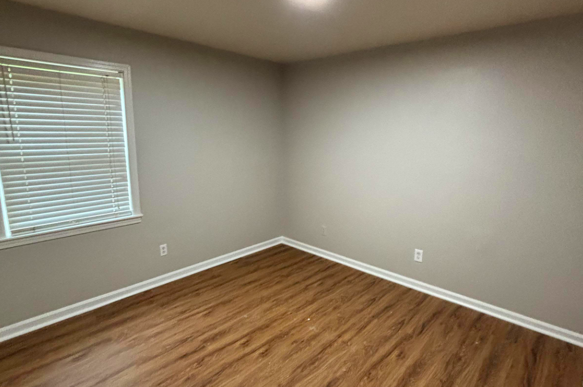 An empty room with light gray walls, a window with horizontal blinds allowing natural light, and a wooden floor. The room has no furniture or decor, emphasizing its spaciousness and minimalism.