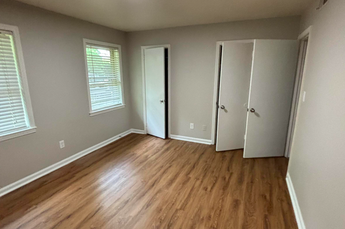A spacious room with light gray walls and hardwood flooring. There are two windows with blinds allowing natural light, and two white doors leading to closets or other areas. The room is empty, creating a clean and open atmosphere suitable for various uses.