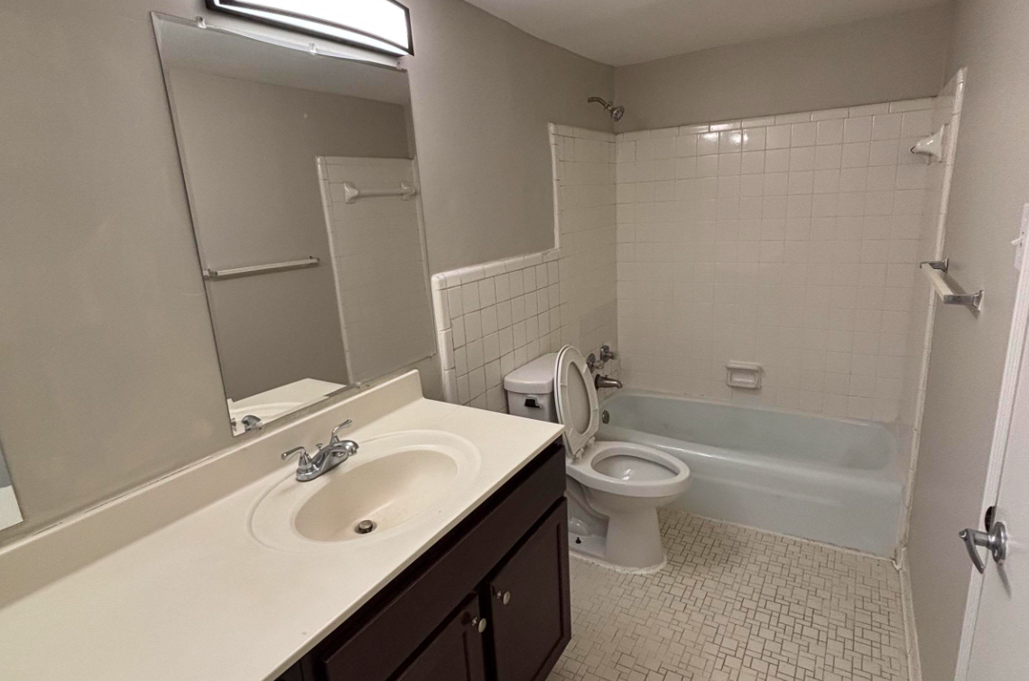 A clean, neutral-toned bathroom featuring a bathtub with a shower, a toilet, and a sink with a dark wooden cabinet. The walls are painted gray, and there is a large mirror above the sink. The floor is tiled in a light-colored pattern.