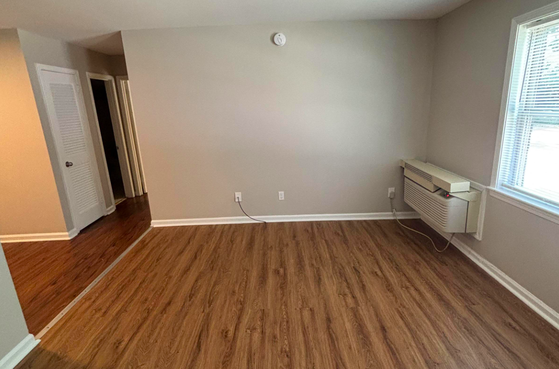 Spacious empty room with light brown laminate flooring, a wall-mounted air conditioning unit, and large windows allowing natural light. Neutral-colored walls lead to an adjacent hallway with doors, creating an airy and inviting atmosphere. Ideal for various interior design possibilities.