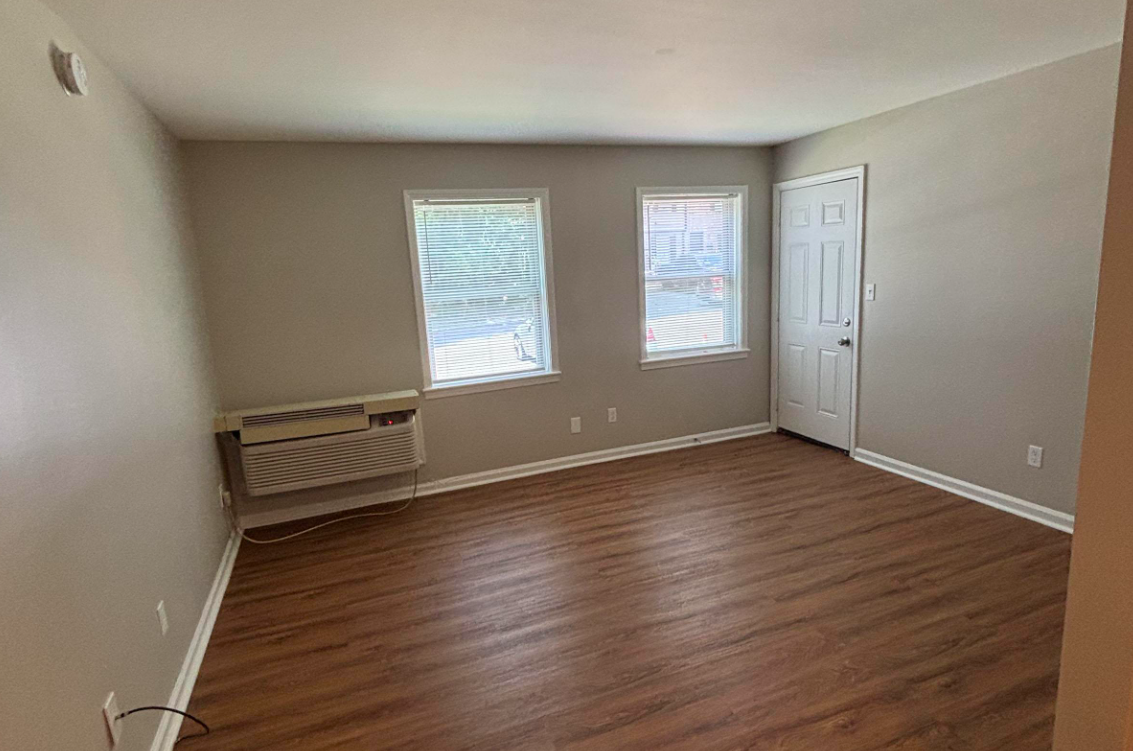 A small empty room with light beige walls and wooden flooring. There are two windows allowing natural light, an air conditioning unit below one window, and a white door on the right. The space appears clean and uncluttered, ideal for potential furniture placement.