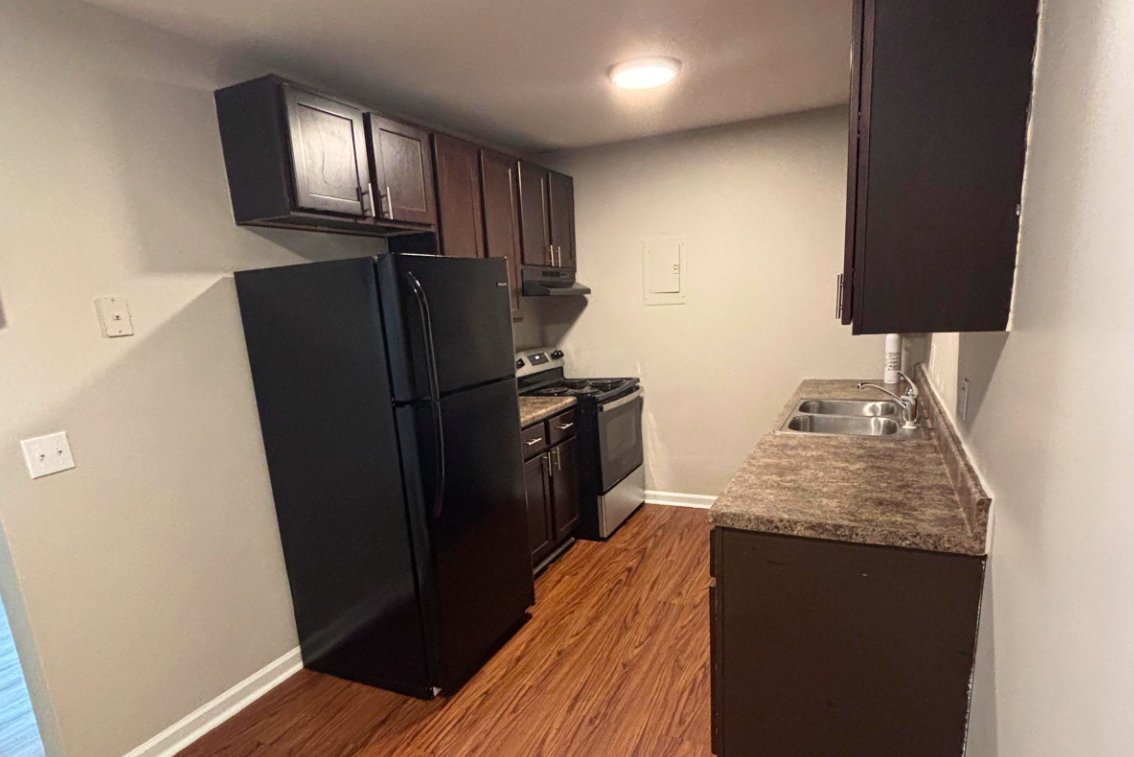 A modern kitchen featuring dark wooden cabinets, a black refrigerator, a stainless steel stove, and a countertop with a double sink. The walls are painted light gray, and the flooring is a warm-toned laminate wood. Bright overhead lighting illuminates the space.