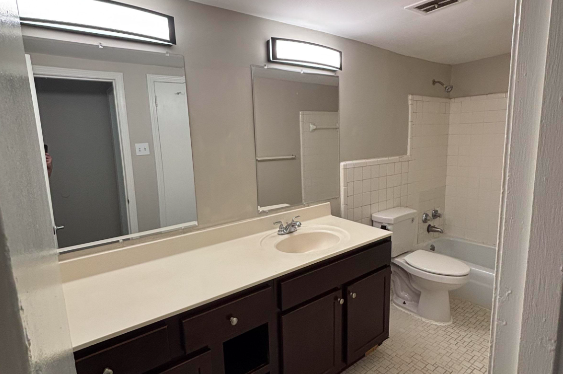 A bathroom featuring a double vanity with two mirrors, a light fixture above, a toilet, and a bathtub with a shower. The walls are painted gray, and the floor has tiled patterns. The cabinetry is dark brown, providing a modern look.