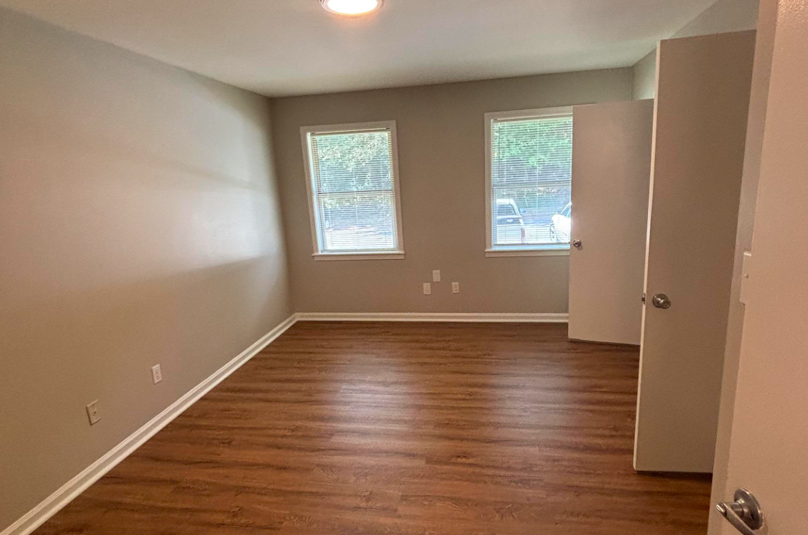 A vacant room featuring light gray walls, two windows allowing natural light, and laminate wood flooring. The room includes a door on the right and has no furniture, creating an open and spacious appearance.