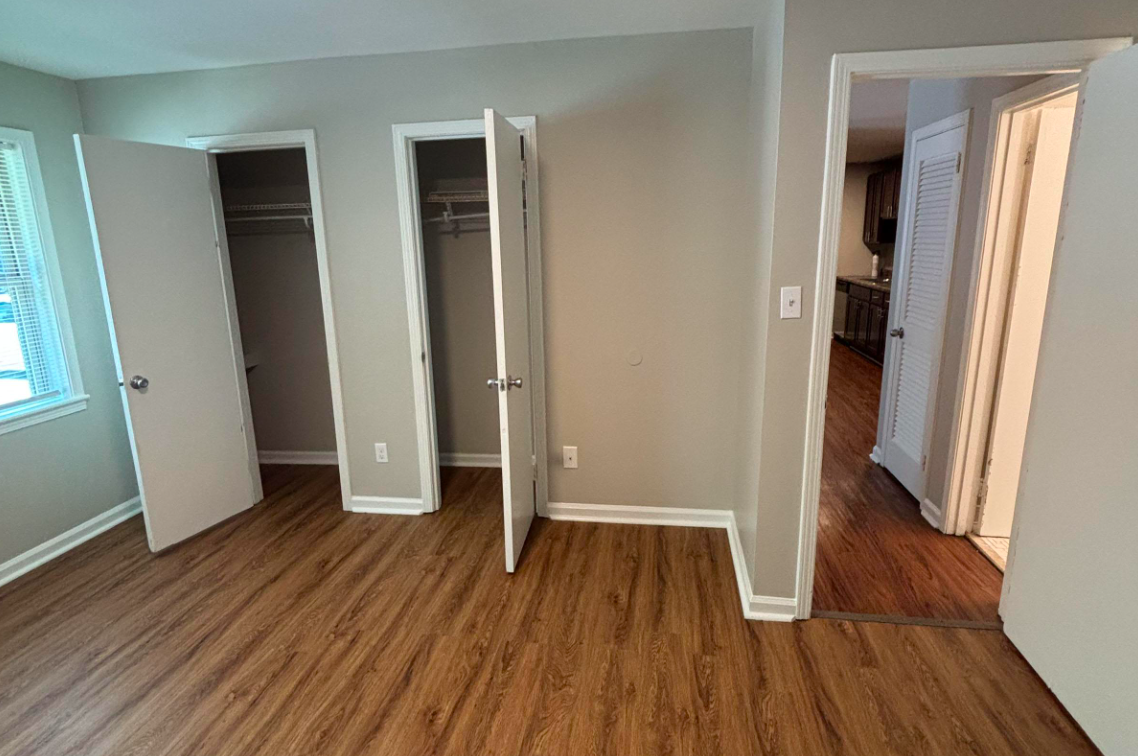 Interior view of a well-lit room featuring two open closet doors, a single closed door to the right leading to another room, and wooden flooring. The walls are painted a light gray, and there are windows on the left providing natural light. The space appears tidy and ready for occupancy.