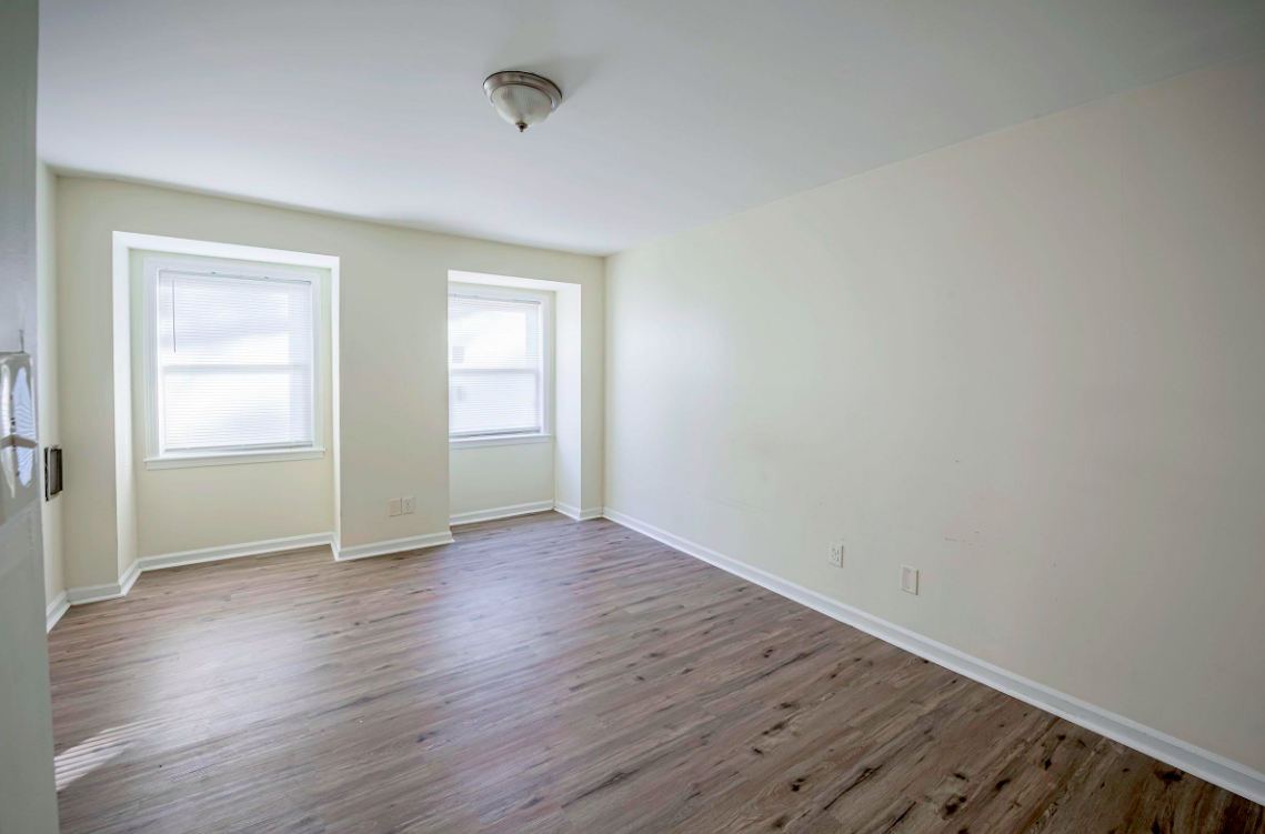 Empty room featuring light-colored walls, two windows with blinds, and hardwood floors. The ceiling has a light fixture, and the room feels spacious and bright.