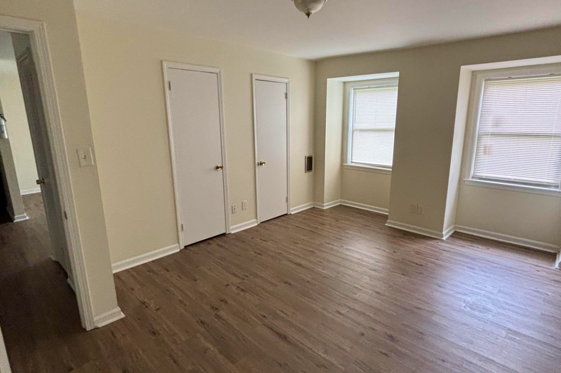 A spacious, empty room with light-colored walls and hardwood flooring. It features two large windows with blinds, providing natural light, and two closed white closet doors. A small wall vent is visible on the left side. The room appears clean and well-maintained.