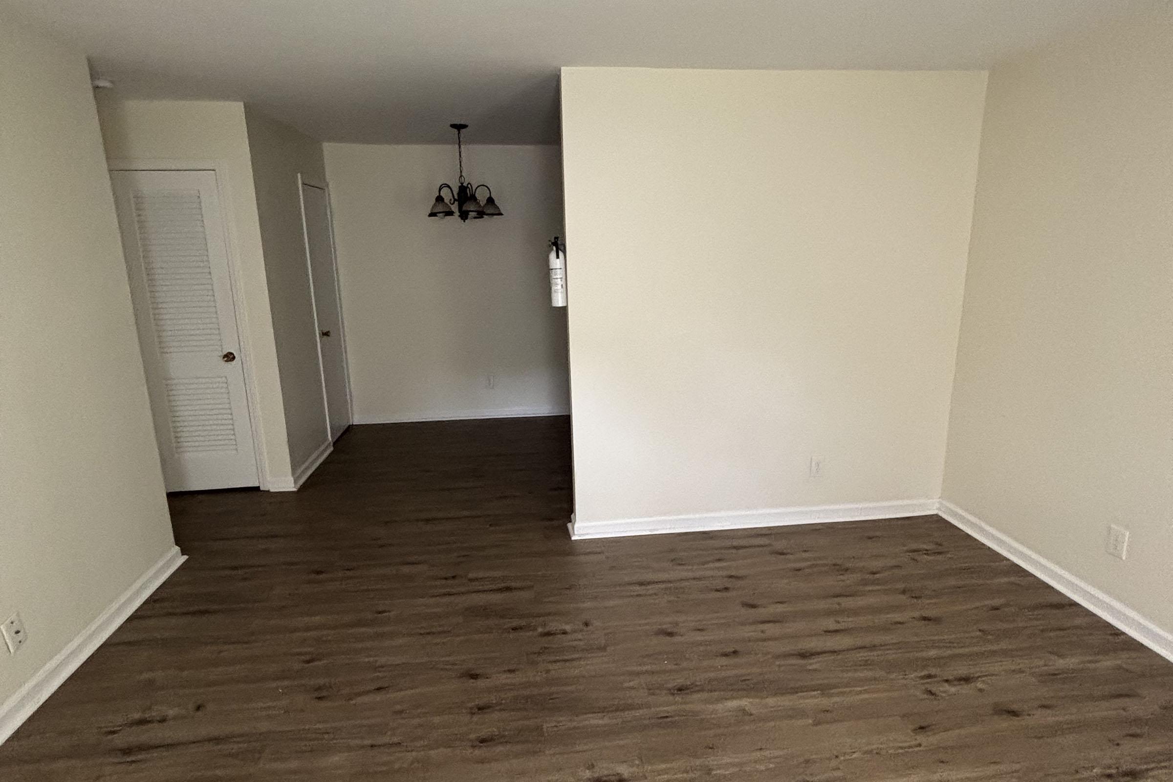 Empty room with light-colored walls and wooden flooring. A doorway is visible on the left leading to a closet, and a ceiling lamp hangs in the center of the space. The layout features an open area, creating a simple and spacious atmosphere.