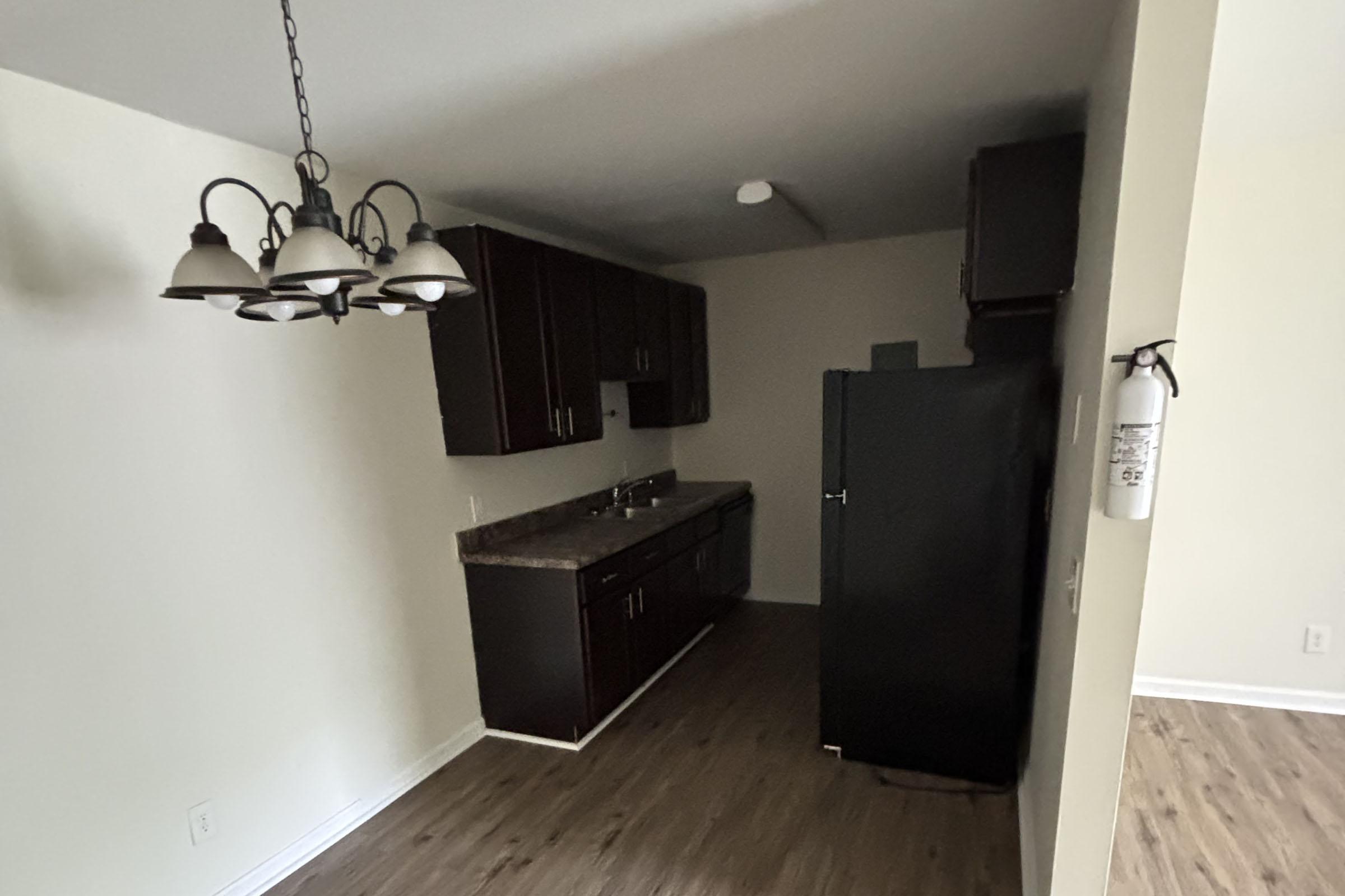 A modern kitchen featuring dark wood cabinets, a black refrigerator, and a countertop. The space has a light-colored wall and wood-style flooring. A chandelier with multiple lights hangs from the ceiling, and a fire extinguisher is mounted on the wall. Natural light brightens the room.