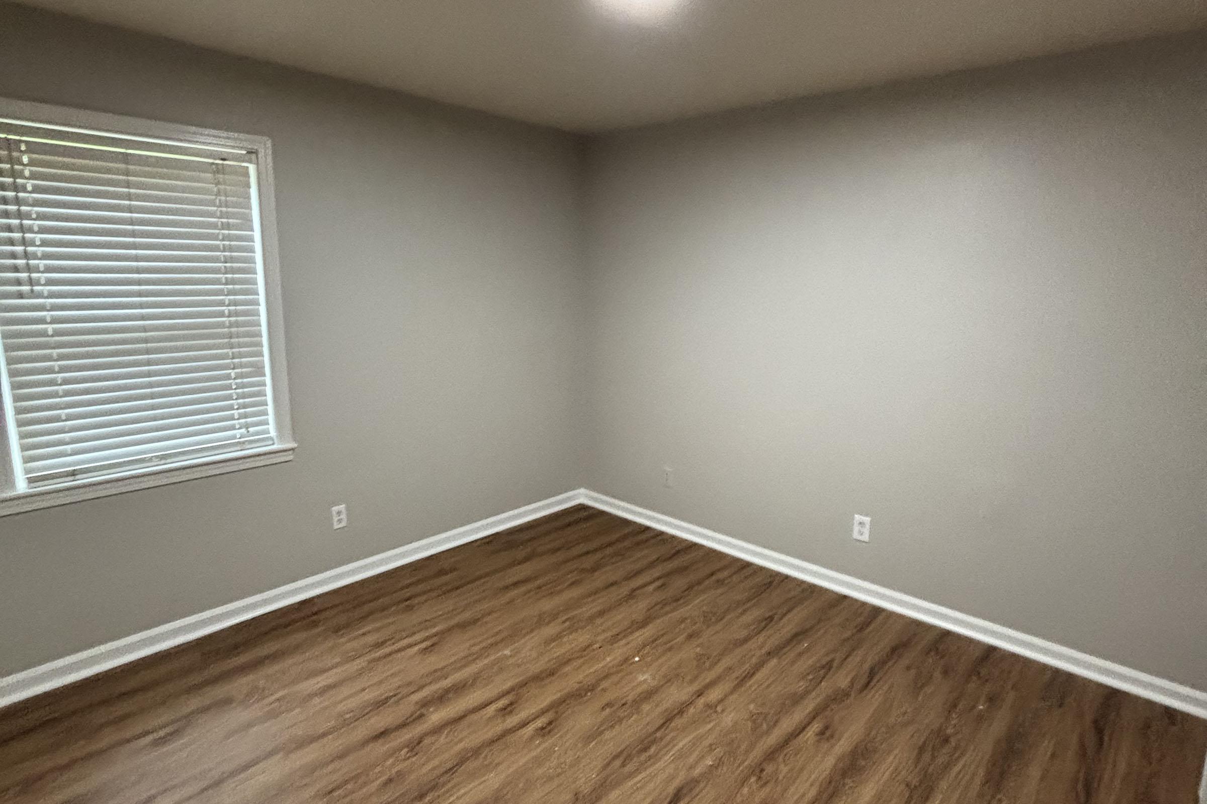 Empty room with light gray walls and a medium brown laminate floor. There is a window on the left with white blinds. The room is unfurnished, creating a spacious and minimalistic appearance. Walls are clean, with no artwork or decorations.