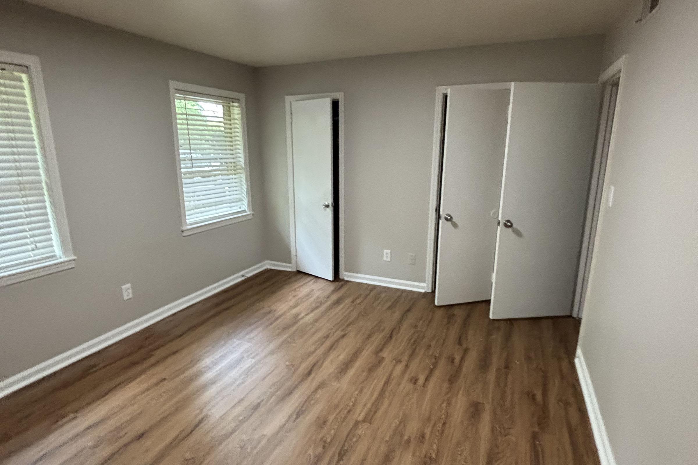 A well-lit room with light gray walls and brown laminate flooring. Two windows with blinds allow natural light in. There are two doors—one closed and one slightly ajar—against the walls. The room appears empty and spacious, suitable for various uses.