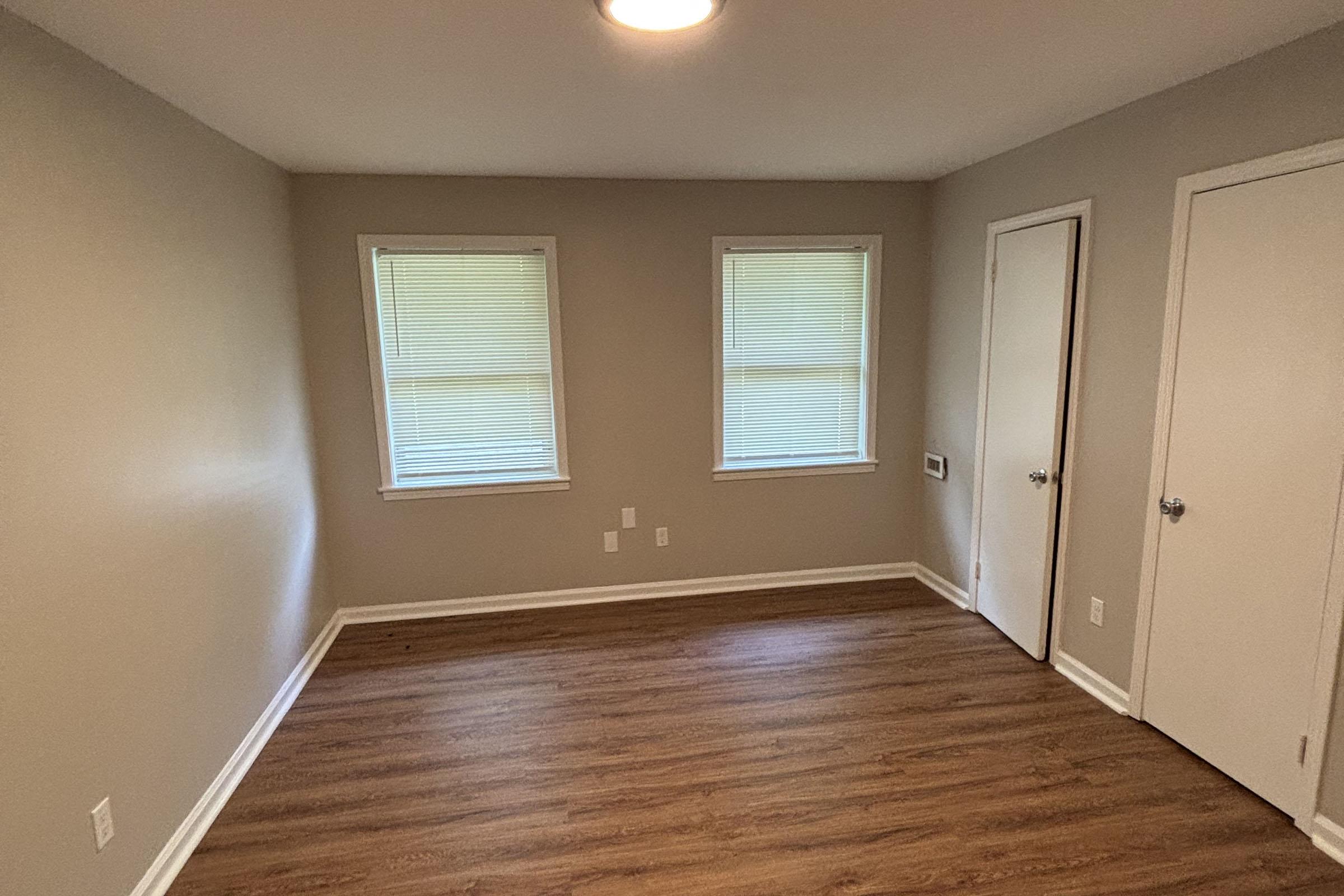 A vacant room with light brown walls and wooden flooring. Two windows with closed blinds allow natural light. There's a closet door on the right and a simple ceiling light fixture. The room is undecorated and appears clean and ready for furnishing.