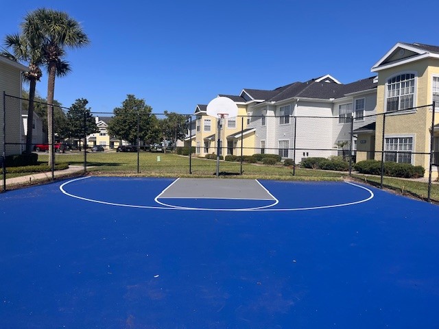 A bright blue basketball court surrounded by a fence, with a white basketball hoop in the center. In the background, there are residential buildings and a clear blue sky, indicating a sunny day. Lush green grass and palm trees are visible, suggesting a pleasant outdoor environment.