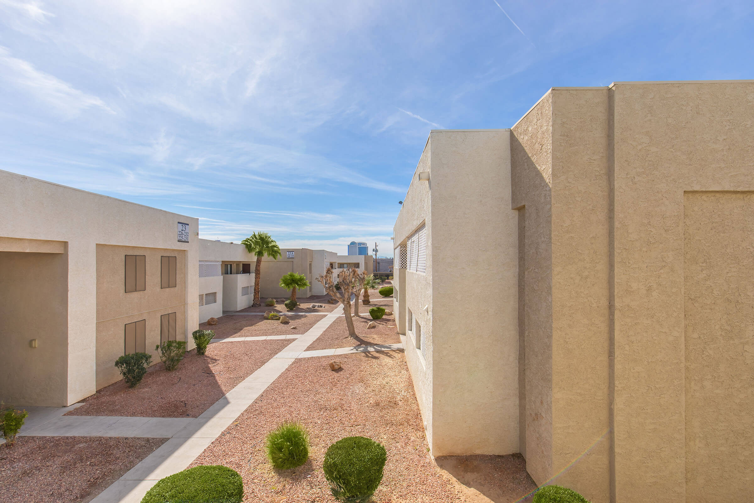 A view of a residential complex featuring beige buildings, landscaped pathways, and small shrubs. The sky is clear with a few clouds, and the background includes distant city skyscrapers, suggesting an urban environment. The scene conveys a sense of tranquility and organization in a desert-like setting.