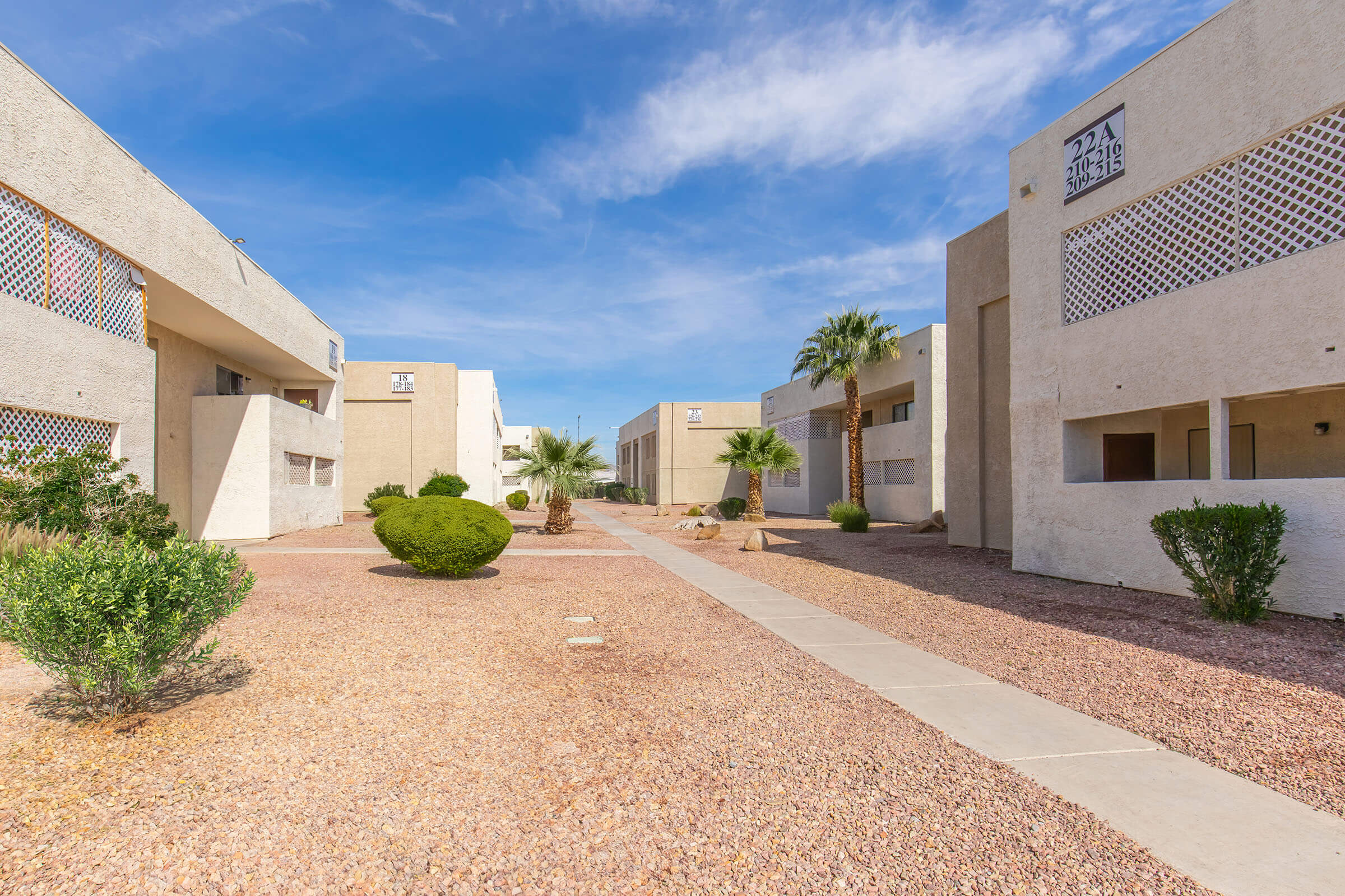 A pathway leading through a landscaped courtyard of a residential complex with several beige buildings, palm trees, and desert plants under a bright blue sky.