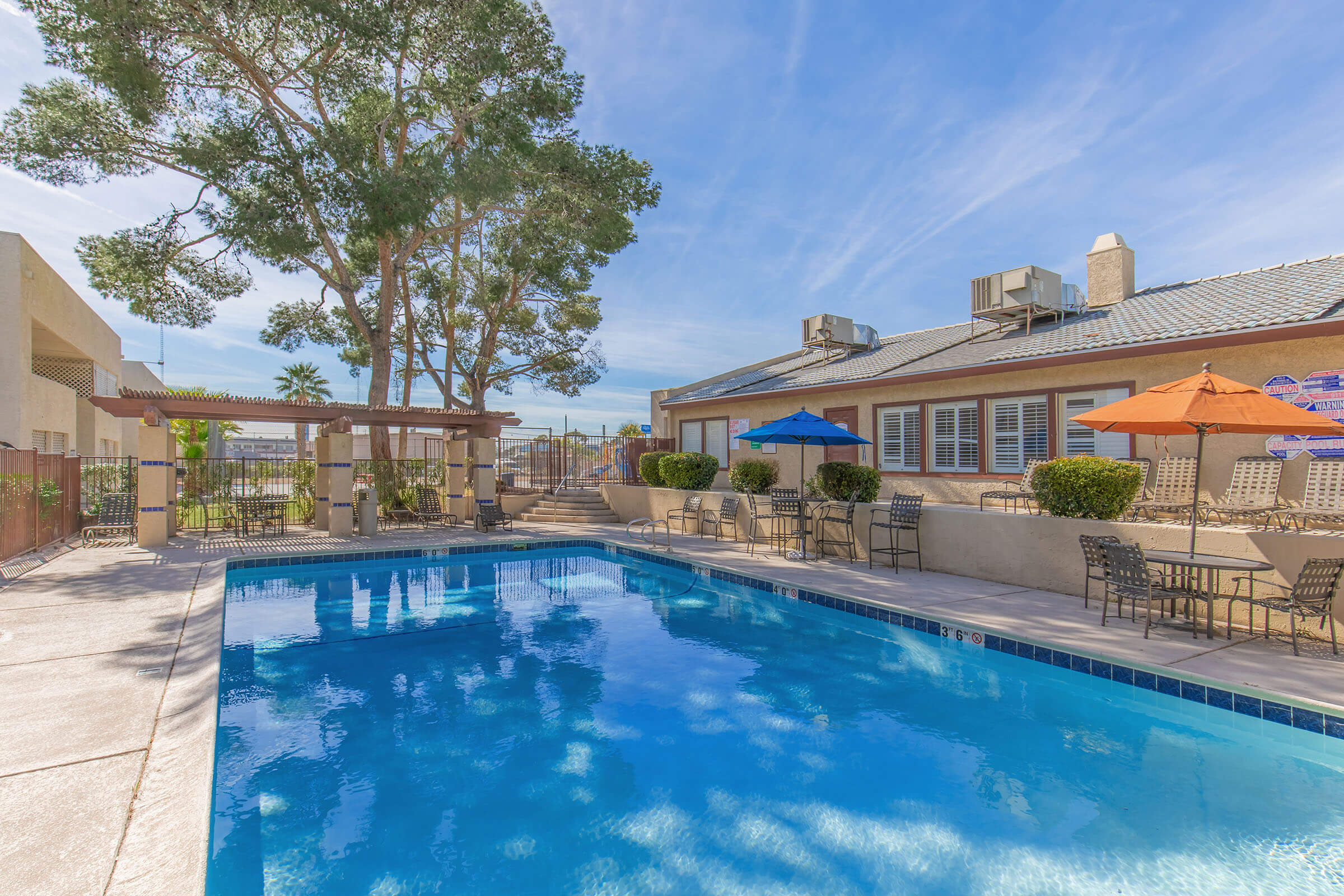 A clear swimming pool surrounded by a patio area with lounge chairs and tables. There are shaded umbrellas in vibrant colors and lush greenery nearby. In the background, a building with large windows is visible, along with a fenced area and a clear blue sky above.