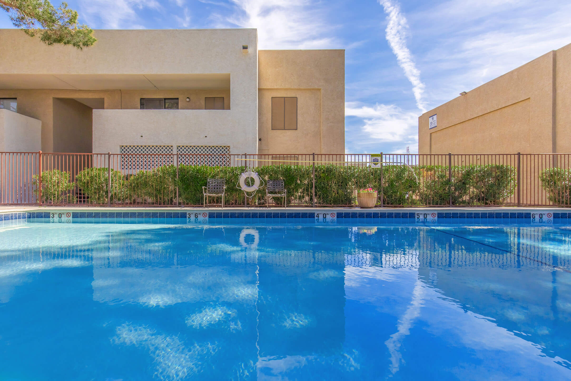 A clear blue swimming pool reflects the sky, surrounded by a fence and lush greenery. In the background, modern building facades are visible under a partly cloudy sky, creating a serene and inviting atmosphere.