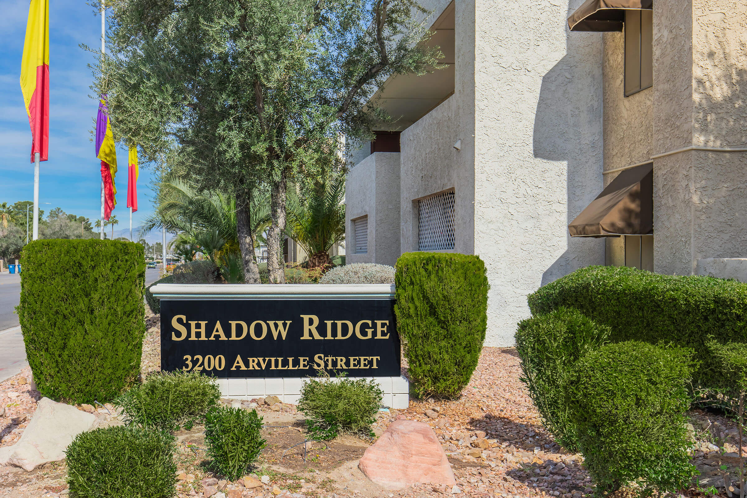 Sign for Shadow Ridge located at 3200 Arville Street, featuring landscaping with trimmed bushes and a pathway. The background includes flags and palm trees, suggesting a welcoming entrance to the property.