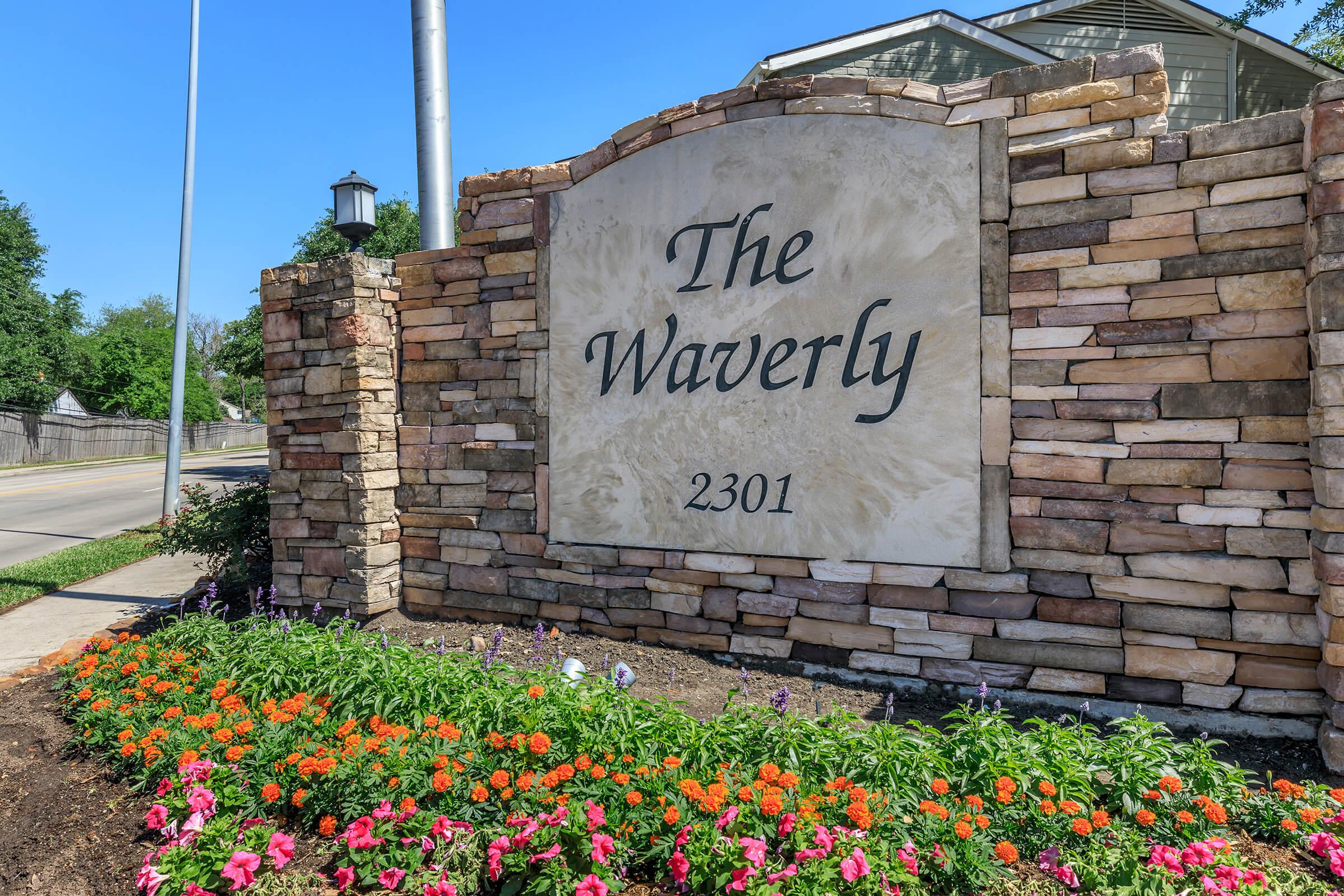 Stone sign for "The Waverly" with the address "2301," surrounded by colorful flowers and greenery. A clear blue sky is visible in the background, and there is a lamp post nearby. The sign features a decorative stone design, giving it a welcoming appearance.