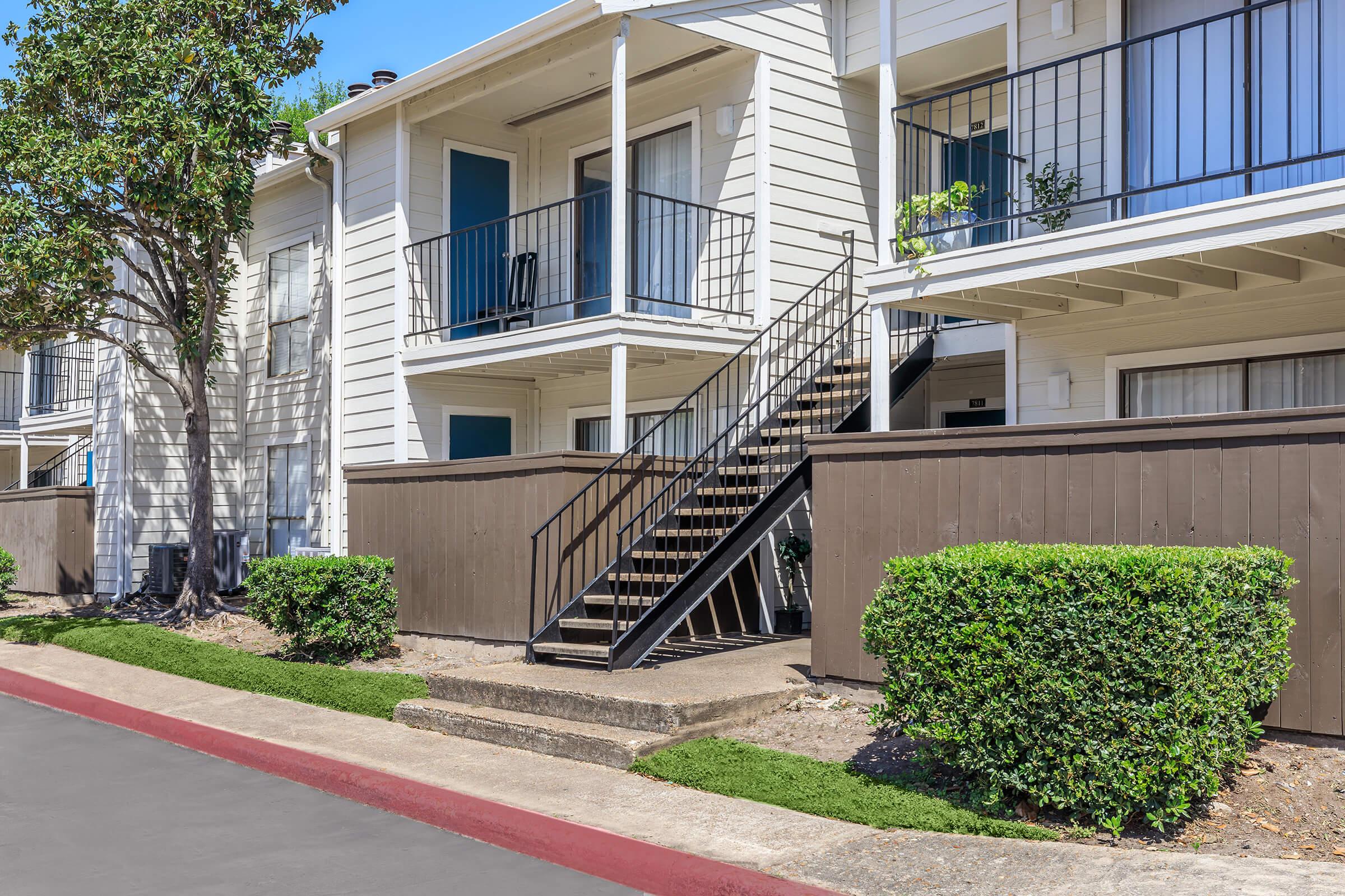 A view of a two-story apartment building with white siding and blue doors. A staircase leads to the second floor, flanked by green bushes on either side. The pathway is paved, and a red curb runs along the side of the building. Sunlight creates a bright atmosphere.