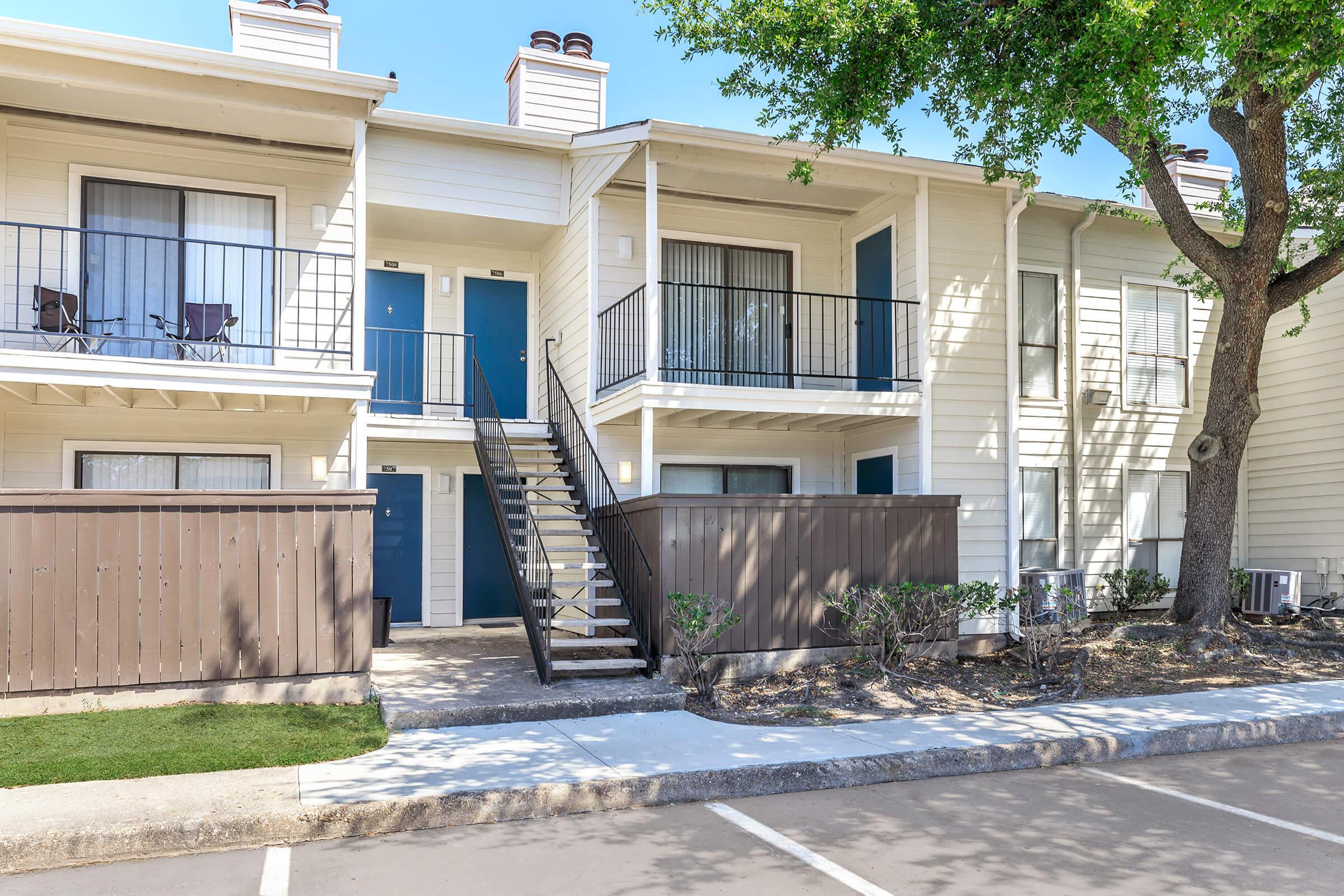 Two-story apartment building with blue doors and white siding. A staircase leads to the upper level, where there are balconies with chairs. The building is surrounded by a small patch of grass, a wooden fence, and a tree. A parking area is visible in front.