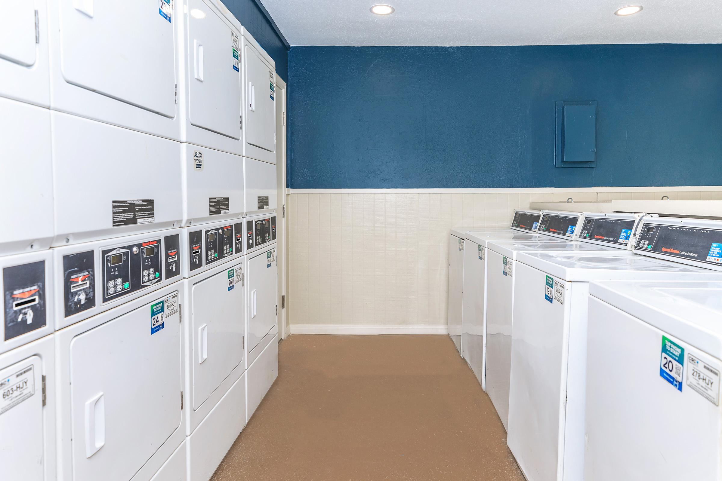 A clean and well-lit laundry room featuring several rows of white washing machines and dryers. The walls are painted blue and there are overhead lights illuminating the space. The floor is finished with a light-colored material, creating a neat and organized environment for laundry activities.