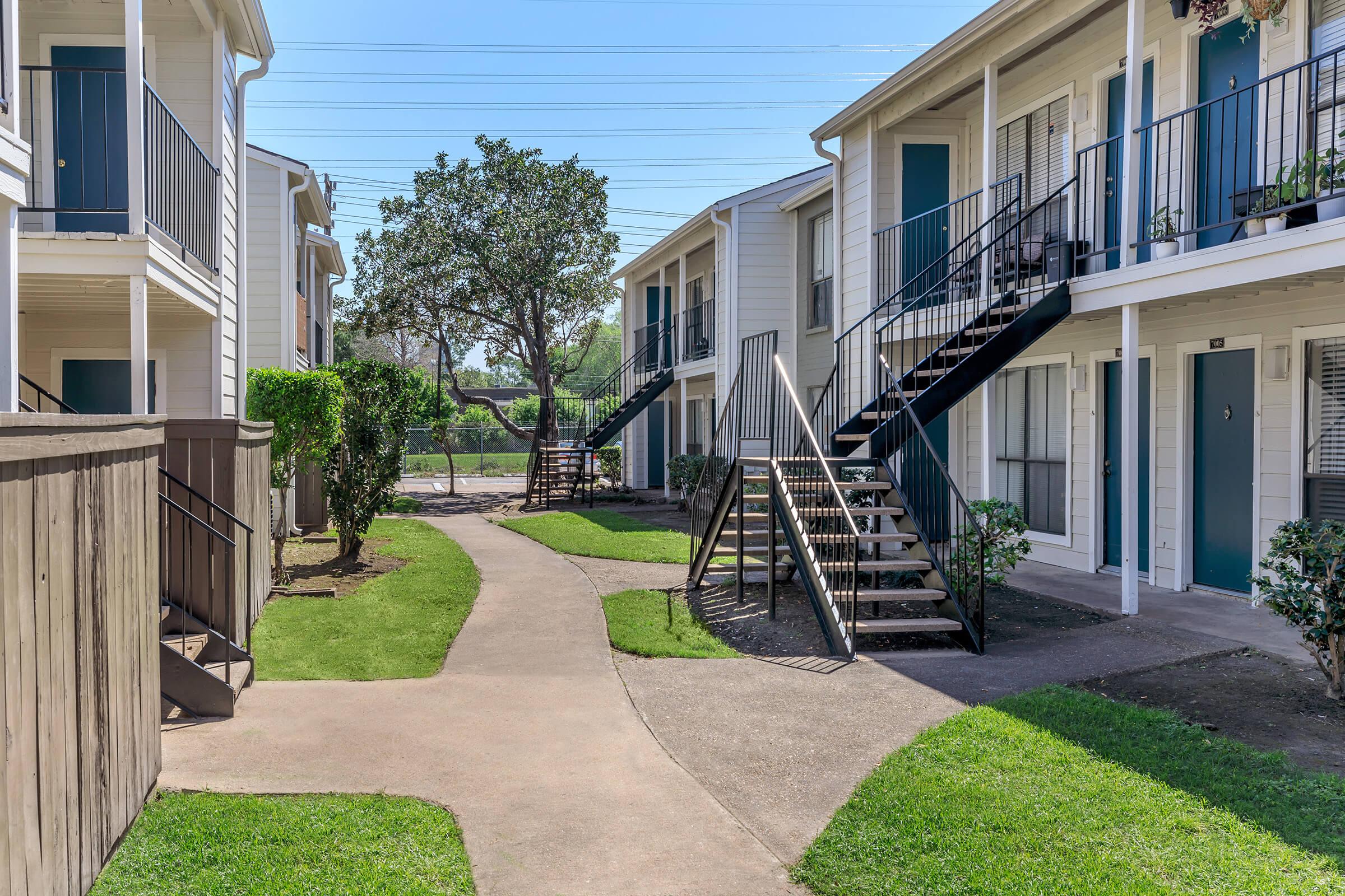A clear view of a residential apartment complex featuring two-story buildings. Each building includes staircases leading to upper entrances. The landscape showcases well-maintained grass, a tree, and a winding pathway connecting the units. Power lines are visible in the background.