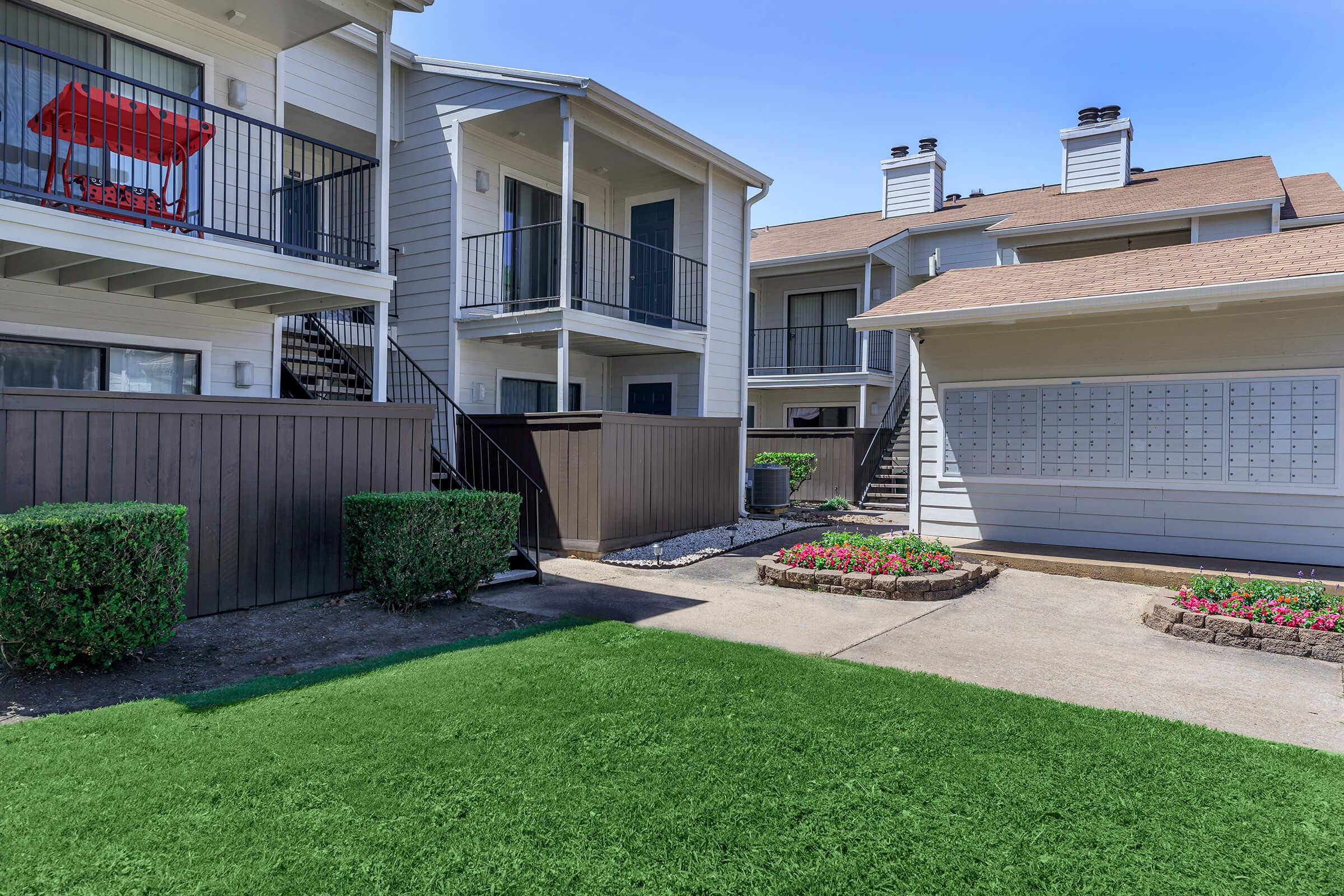 View of an apartment complex featuring two buildings with balconies, a green lawn, and flower beds. Mailboxes are visible along the side of one building, and there are bushes and a pathway between the two structures, set against a clear blue sky.