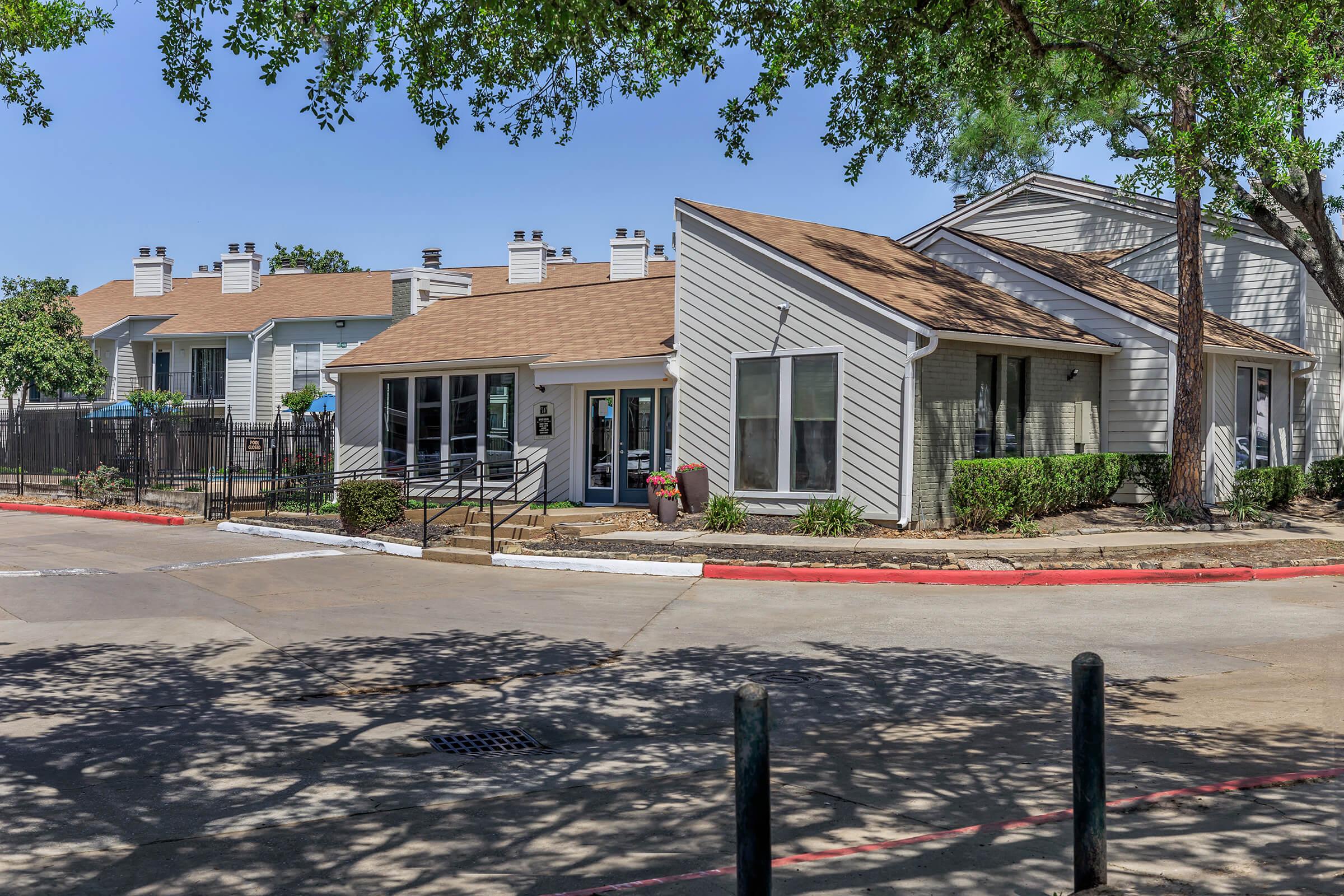 A sunny residential complex features a modern building with a sloping roof, surrounded by well-maintained landscaping. The background showcases other units and a paved road leading toward the entrance. The scene reflects a welcoming atmosphere with clear blue skies.